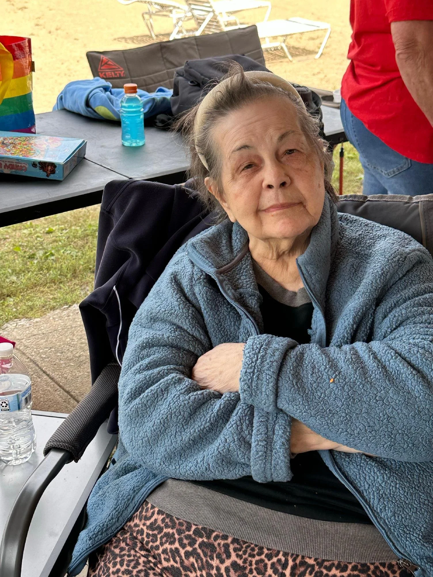 An elderly woman wearing a light blue zip-up jacket and a beige headband is sitting outside at a table, smiling with eyes partially closed. The table has a bottle of water, a colorful bag, some clothing, a backpack, and a board game on it. There are 