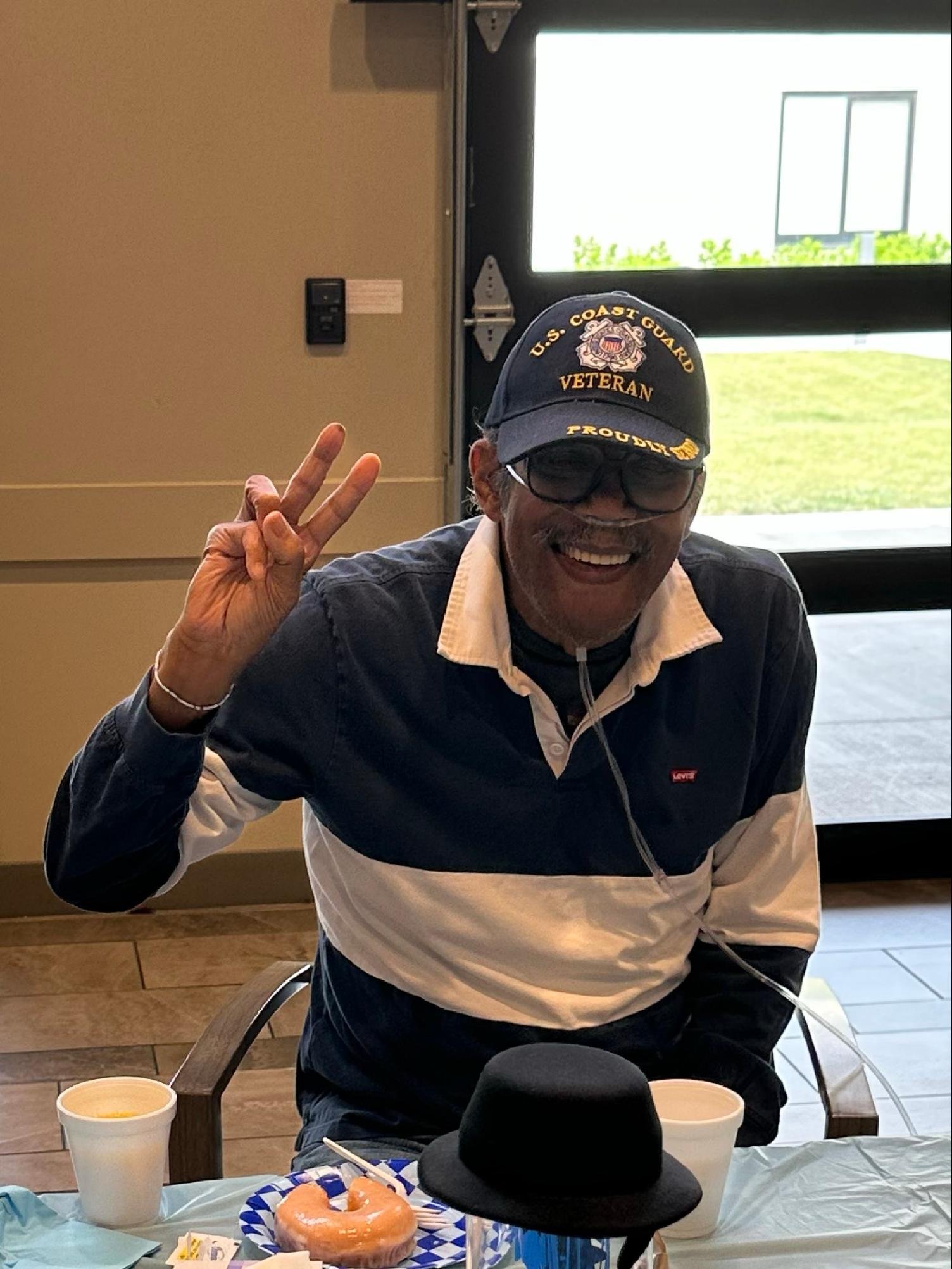 A man smiling and making a peace sign with his fingers, wearing a U.S. Coast Guard veteran cap with sunglasses on top, sitting at a table with a donut, two cups of soup, and a black hat. The background shows a door with windows and bright outdoor sce