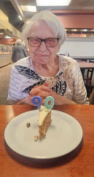An elderly woman with white hair and glasses sitting at a table in a restaurant, celebrating her 90th birthday with a slice of cake with blue and pink number candles reading '90'.