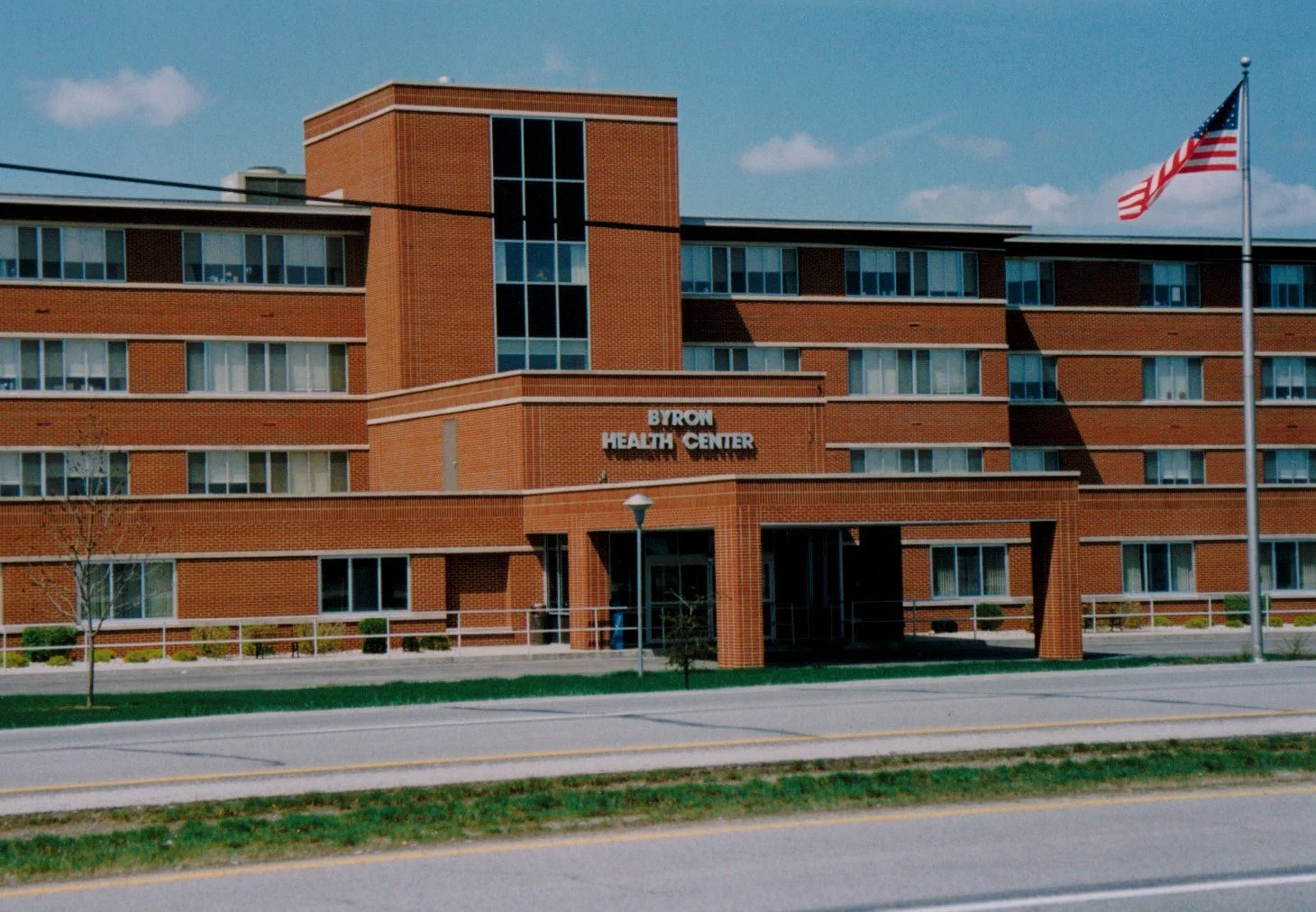 Front view of Byron Health Center, a multi-story brick building with several windows, located along a street with a sidewalk, streetlamp, and an American flag flying on a flagpole.