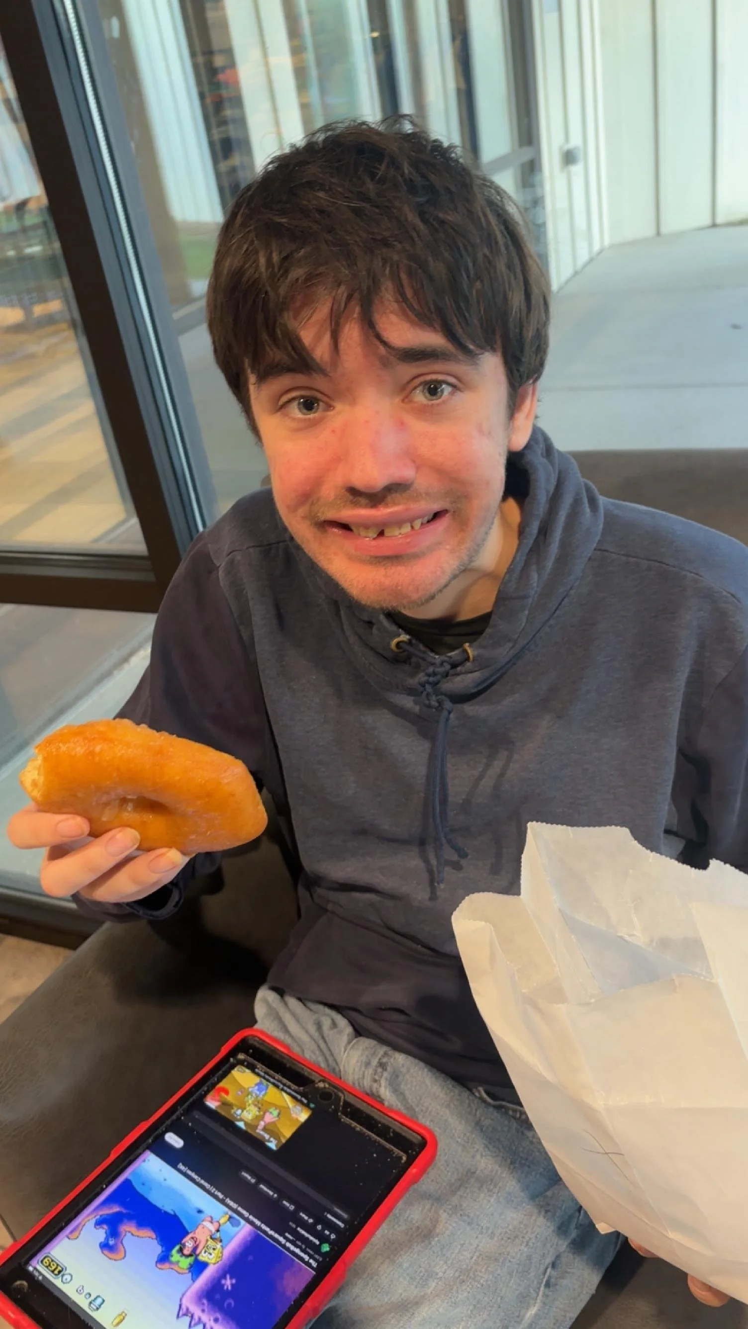 A young man with dark, messy hair and light skin smiling and holding a half-eaten donut in his right hand, sitting indoors near large windows. A smartphone displaying a colorful game is on his lap, and there is a white paper bag in his left hand.