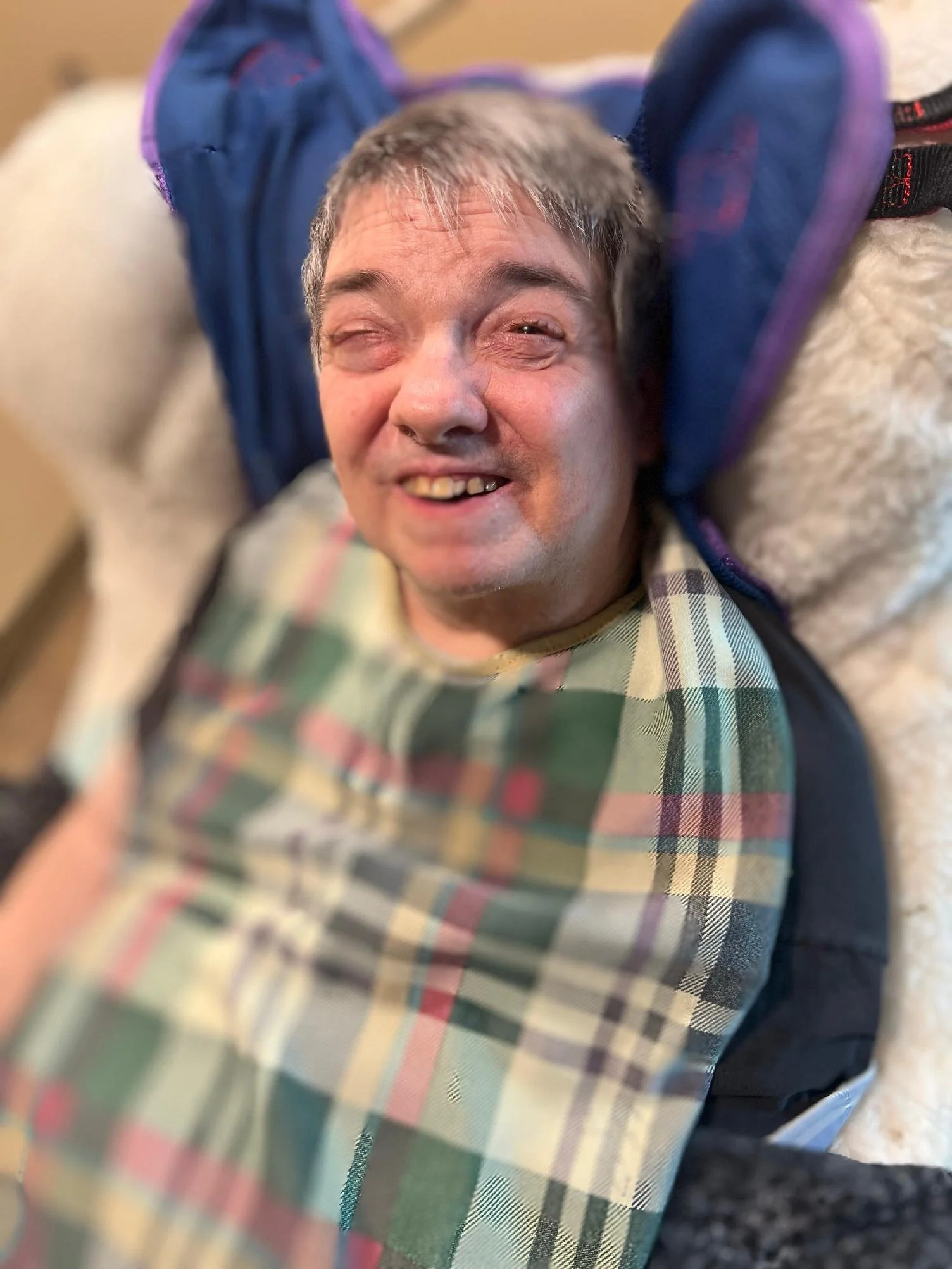 A person lying on their back on a soft surface, smiling at the camera with their head near a plaid cloth. They are wearing a dark jacket and a plaid shirt. The background is slightly blurred.
