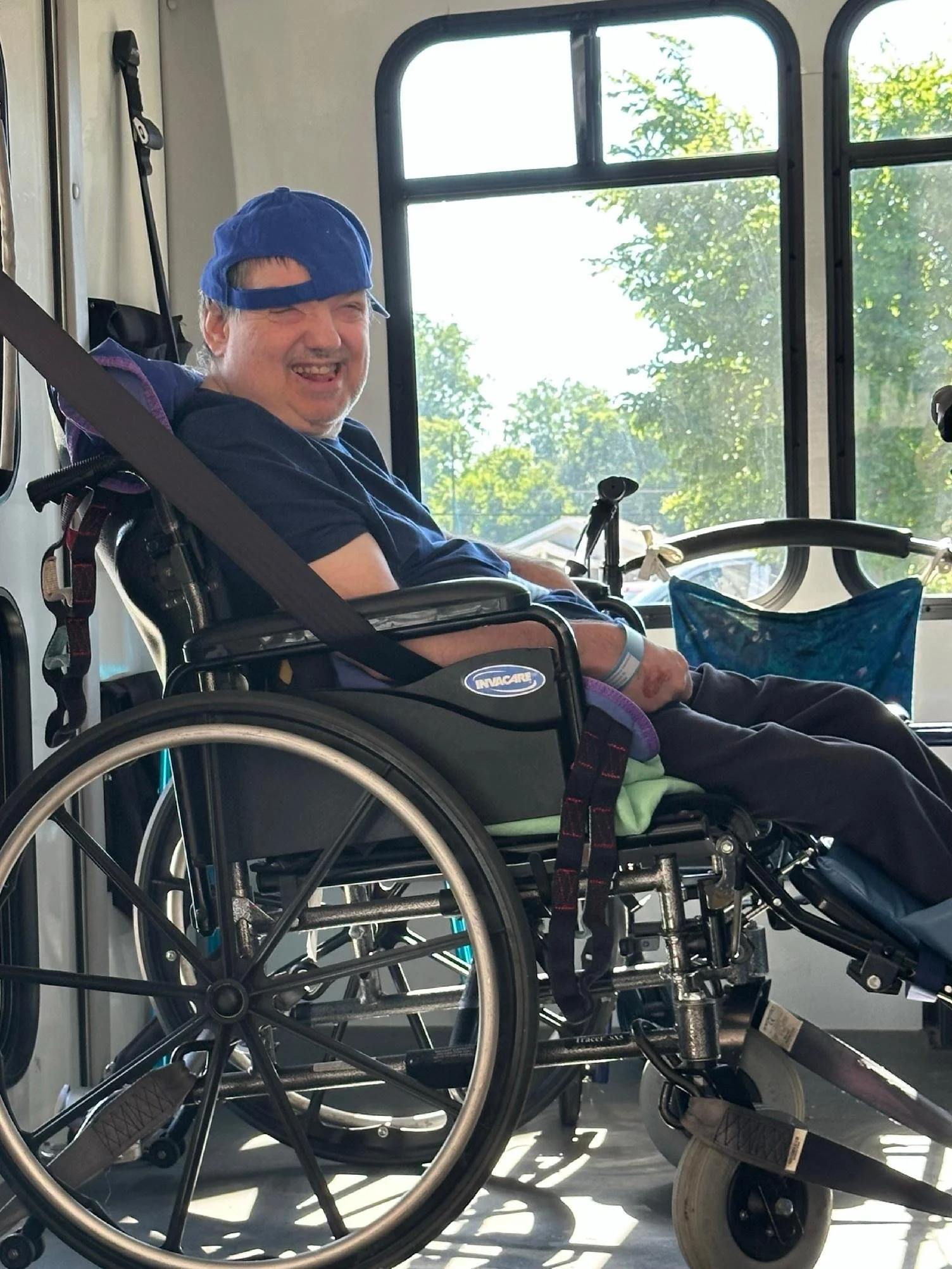 A man sitting in a wheelchair inside a vehicle, smiling and wearing a blue cap. Large windows with a view of trees outside.