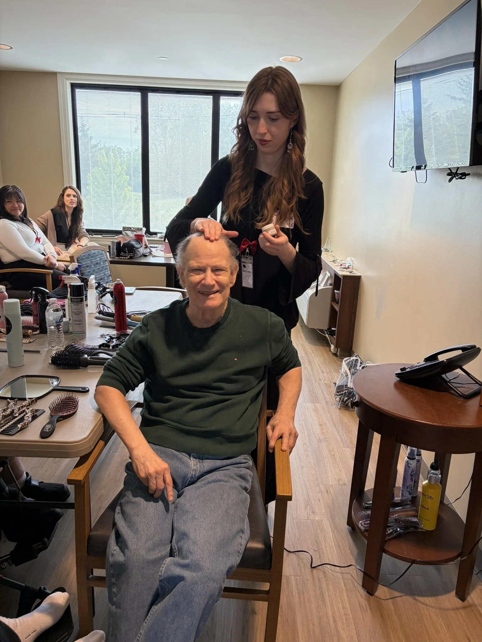 A woman receiving makeup or hair styling from a professional in a room with other people sitting at tables and a large window in the background.