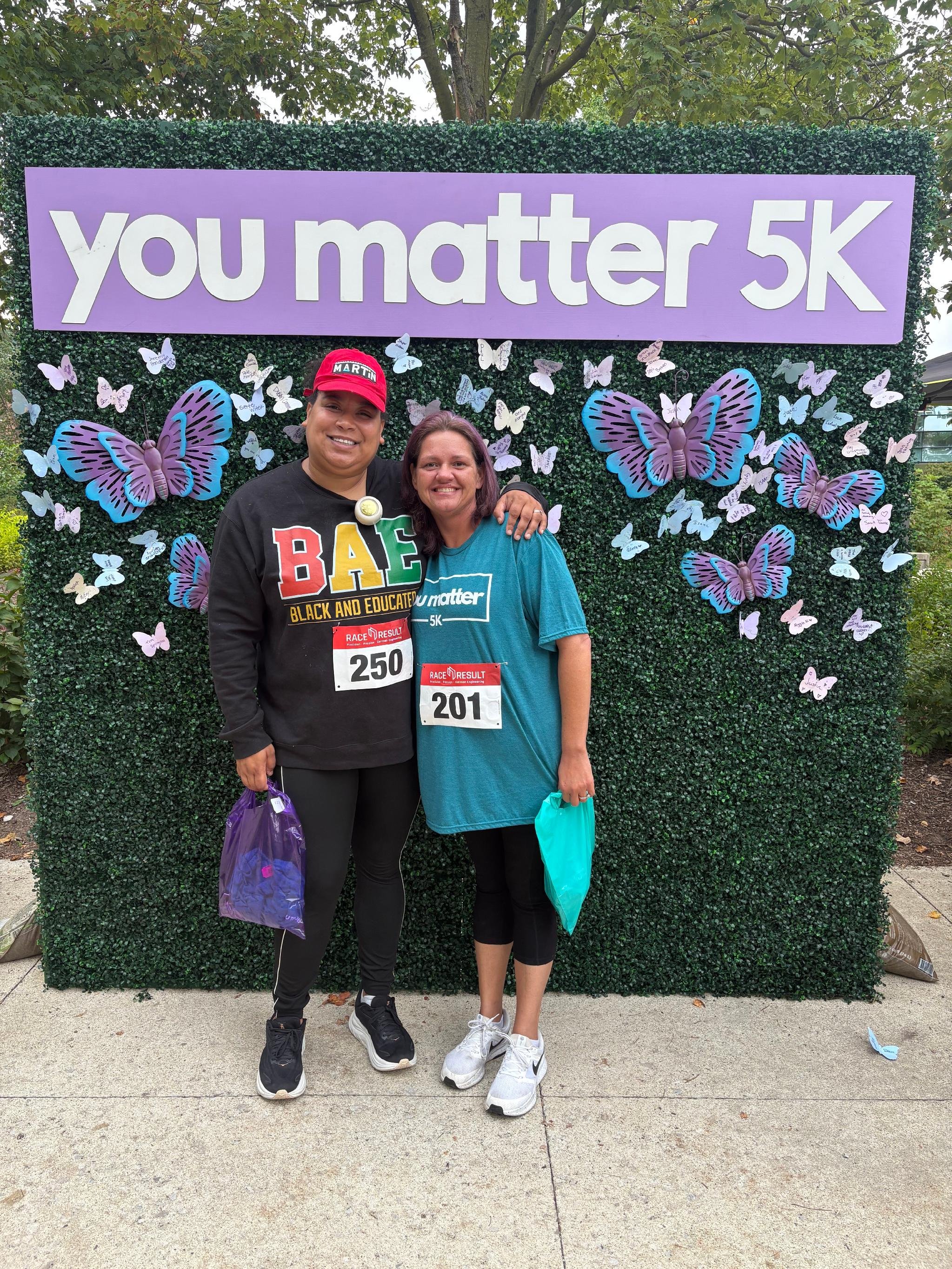 Two women at a 5K race event standing in front of a purple sign that says 'you matter 5K' surrounded by purple and white butterfly decorations. They are smiling, wearing race bibs, and holding bags, with a green hedge and trees in the background.