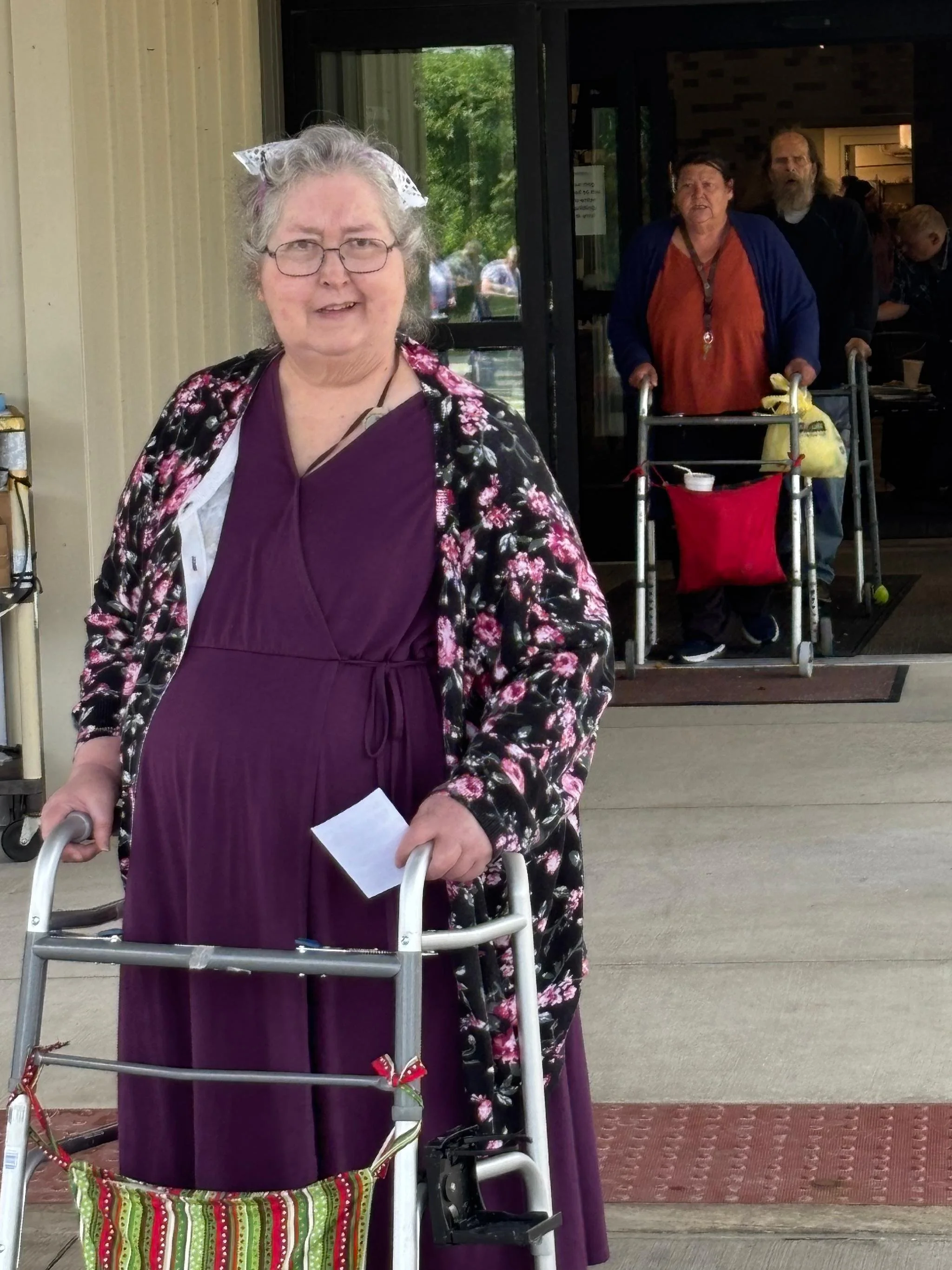 An elderly woman with glasses smiling, holding a walker, in front of a building entrance. Behind her, two people with walkers and a man with a beard are visible inside the building.