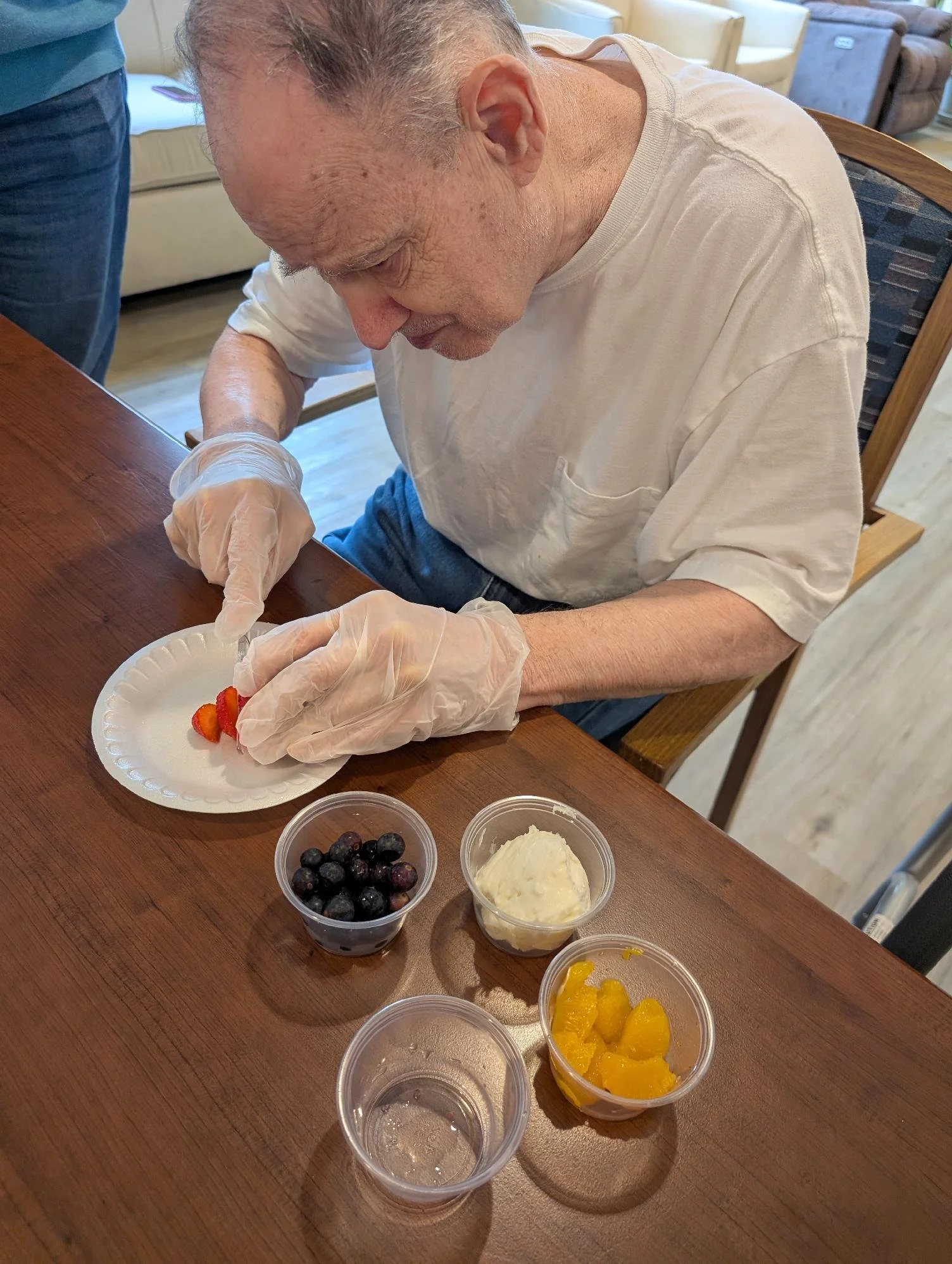 An elderly man with gray hair wearing a white T-shirt and gloves is assembling or serving dessert on a paper plate. There are containers of blueberries, whipped cream, and orange slices on the table.