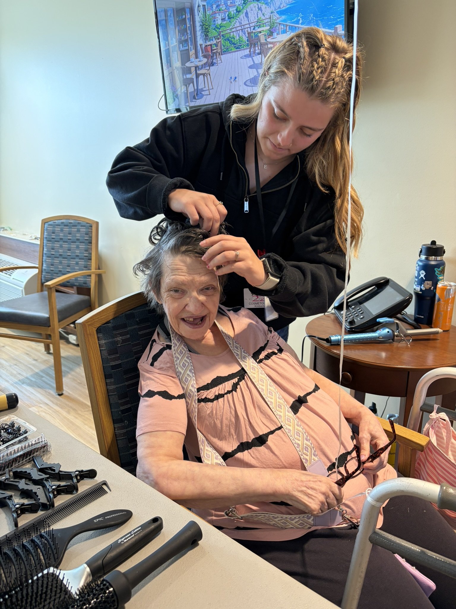 An elderly woman sitting in a chair getting her hair done by a young hairstylist. The woman is holding glasses and smiling. The hairstylist is working on her hair, which is styled with curls. There are hairdressing tools and items on the table in the