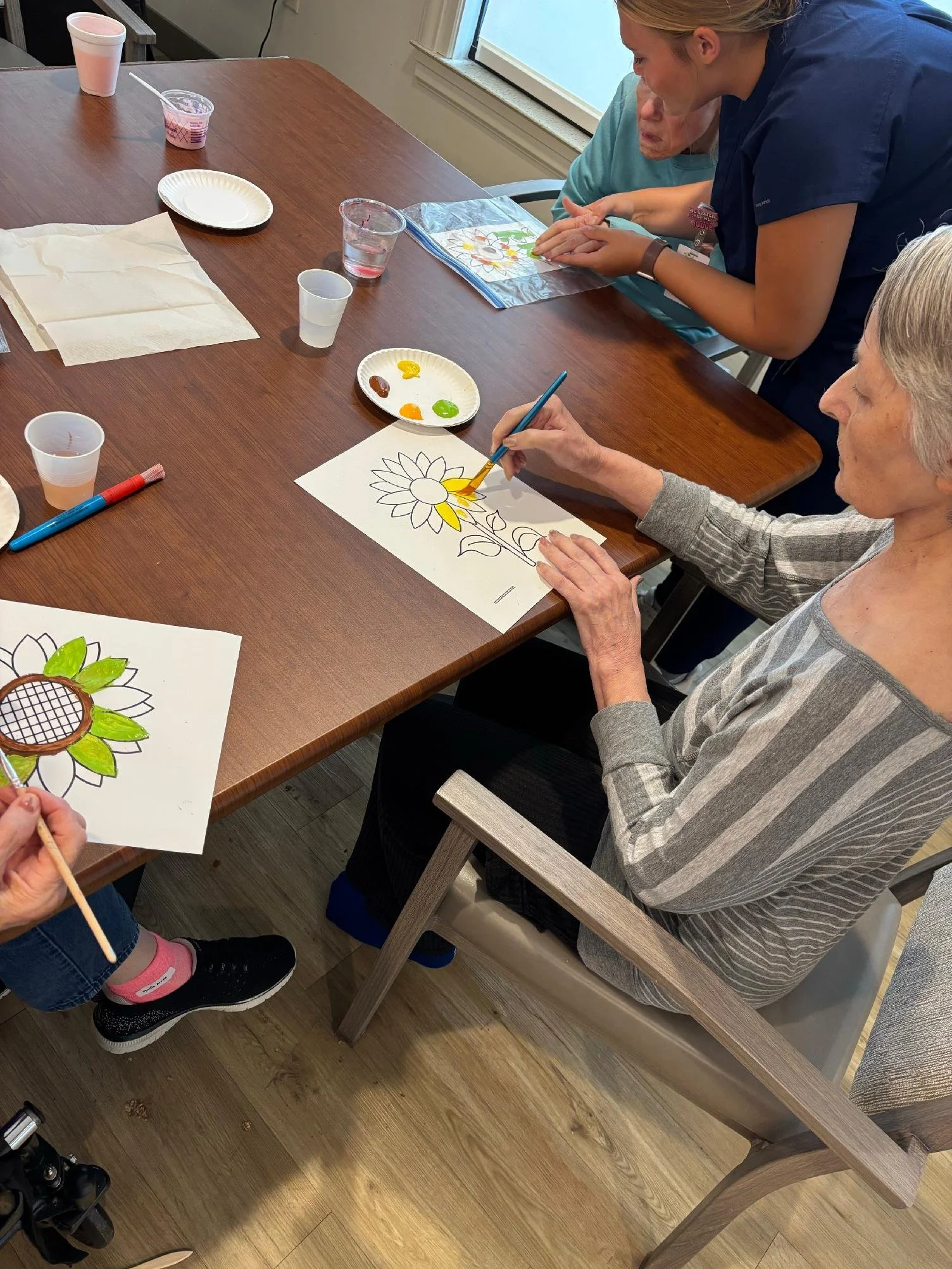 Older woman painting a sunflower on paper, with a younger woman helping, in a room with a wooden table and various art supplies.