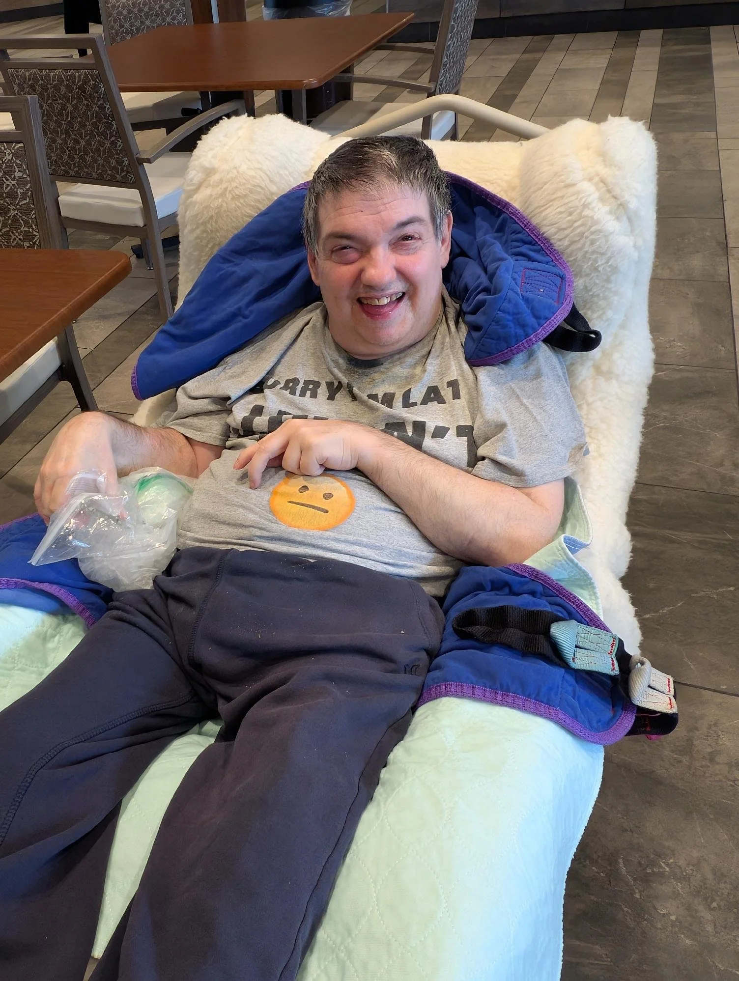 Man with short dark hair and smiling face reclining in a wheelchair, wearing a gray t-shirt and dark pants, with a purple and blue blanket over his shoulders, in a restaurant or dining area with wooden tables and chairs.