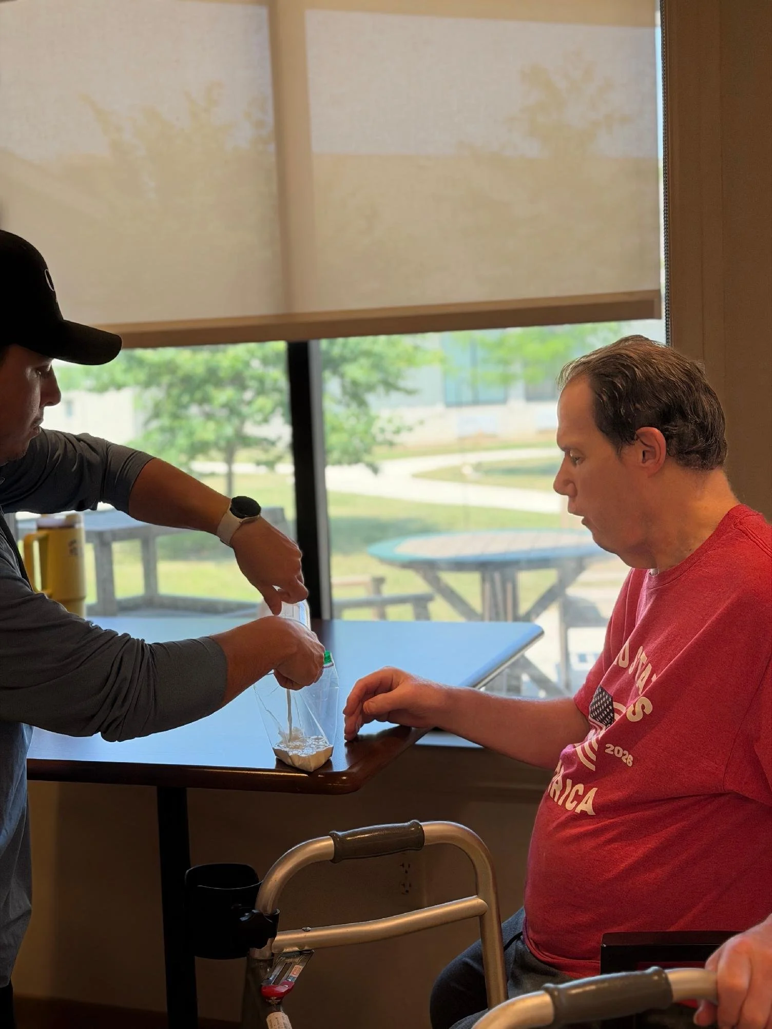 A man in a red shirt sitting at a table with a walker in front of him, receiving a small plastic bag from another person, inside a room with large windows and a view of outdoor tables and greenery.