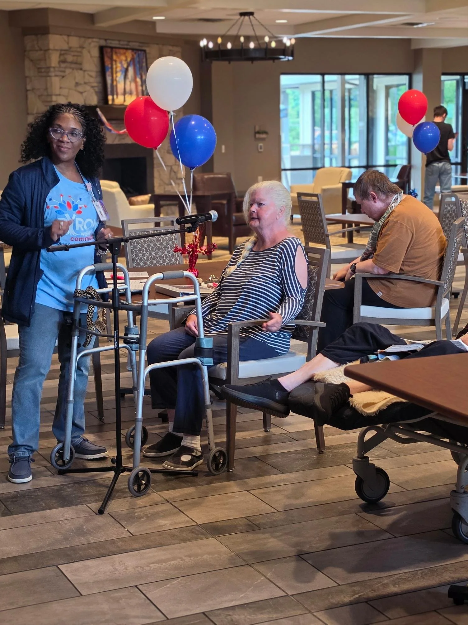 An indoor scene with elderly individuals at a gathering, some sitting and some standing, with balloons in red, white, and blue. One woman with white hair is sitting in a chair with a walker in front of her, and another woman with curly hair and glass
