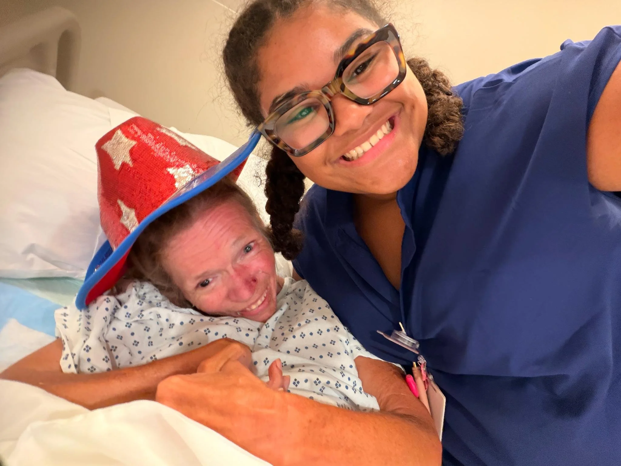 A woman lying in a hospital bed with an IV and hospital gown, smiling at the camera, alongside a young girl wearing glasses and a hospital gown, also smiling and taking a selfie with her.
