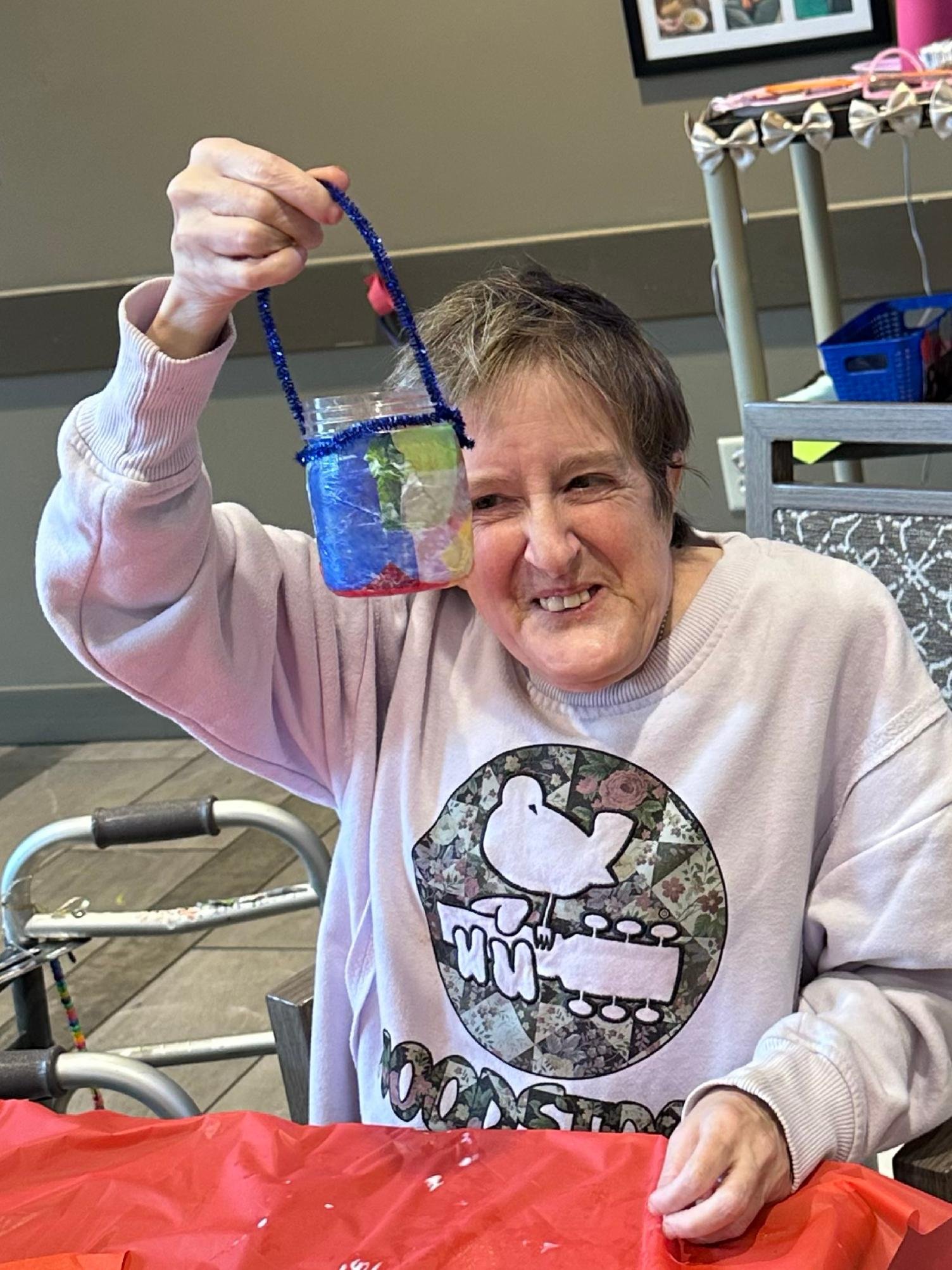 An elderly woman with short gray hair smiling, holding a blue pipe cleaner. She is wearing a pink sweatshirt with a graphic print on the front. The setting appears to be indoors with wall art and shelves in the background, and a walker is visible bes