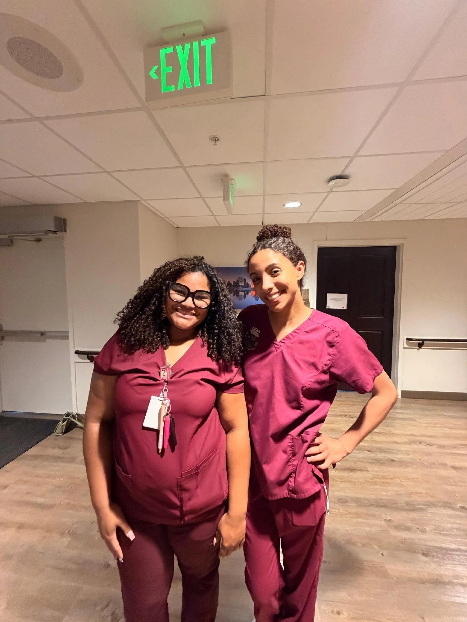 Two women in burgundy scrubs pose together indoors, smiling at the camera, with an exit sign illuminated in green above and a door in the background.