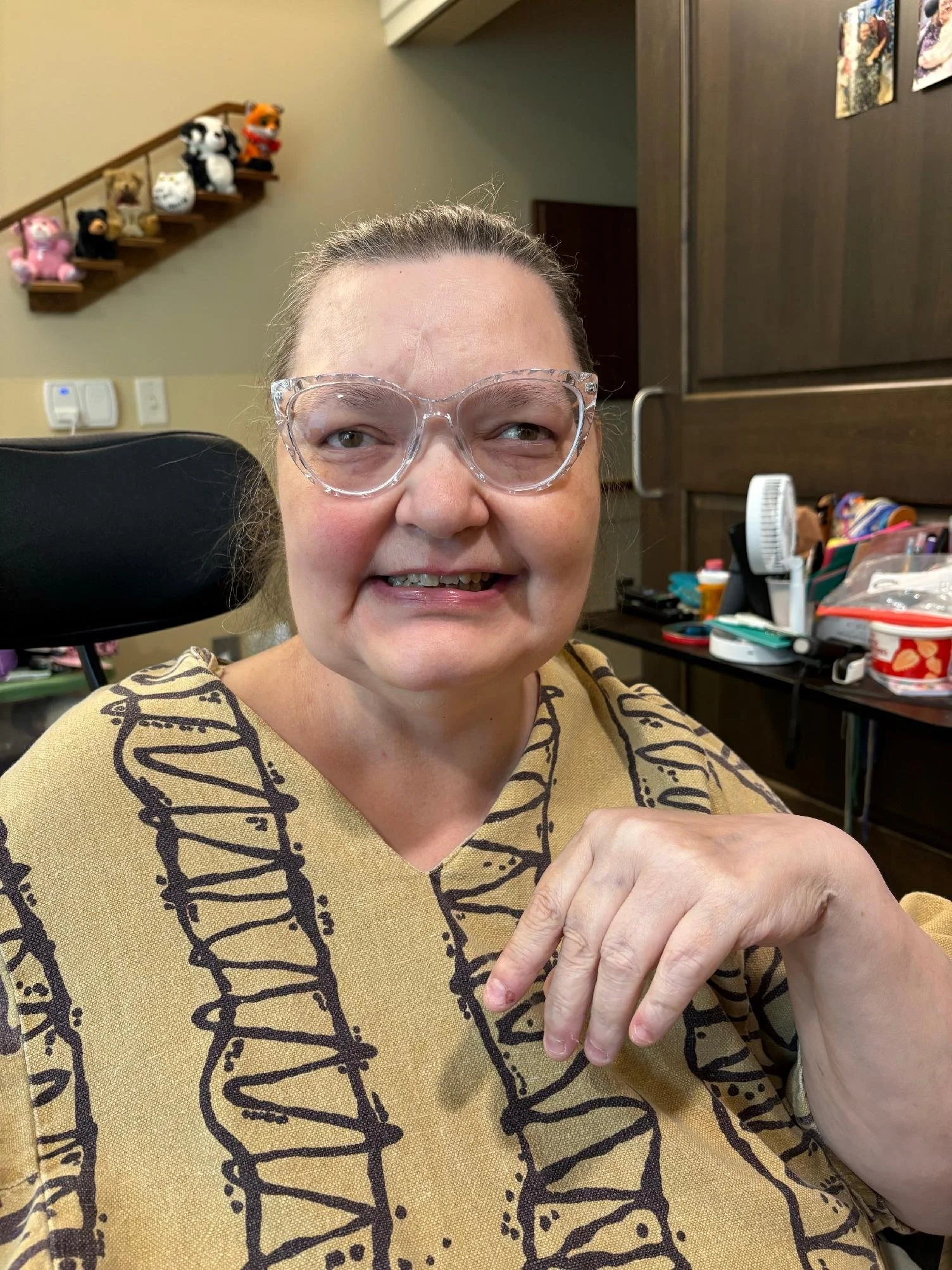 A middle-aged woman with glasses smiling, sitting in a room with a collection of plush toys on a staircase and various items on a desk in the background.