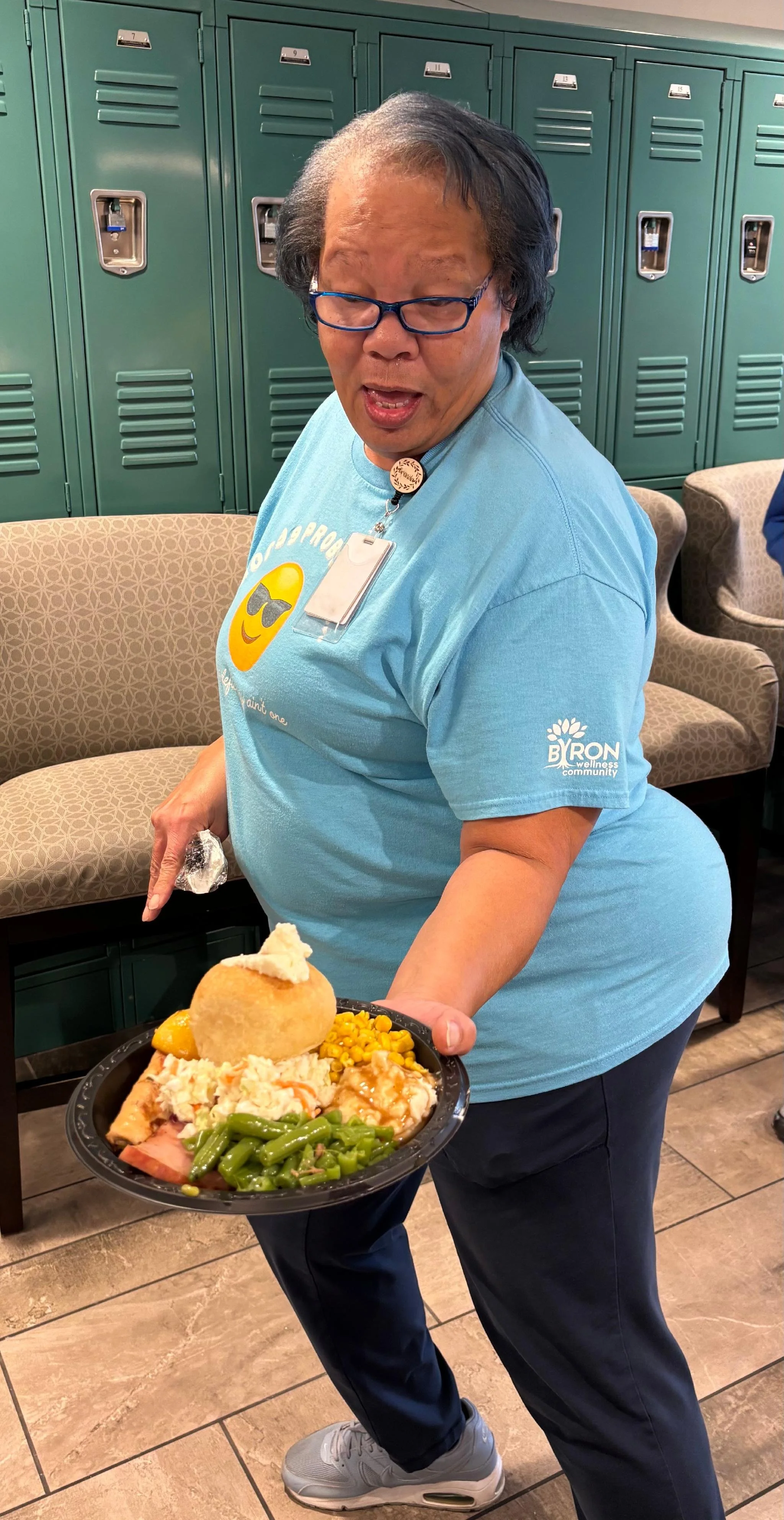 A woman wearing glasses and a blue t-shirt with 'BYRON Wellness Community' logo holds a plate with various food items, including green beans, macaroni and cheese, a breaded item, and a scoop of mashed potatoes with gravy, standing in front of teal lockers.