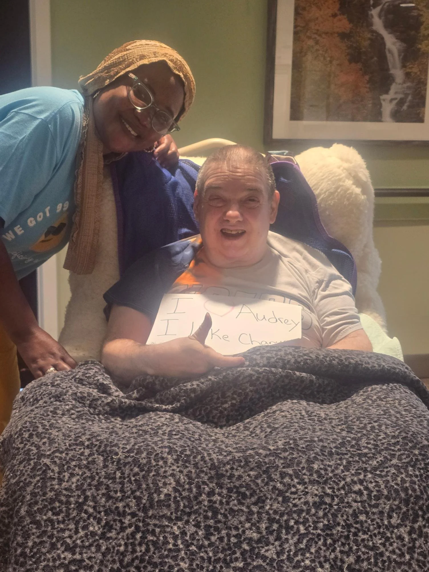 A man in a wheelchair holding a handwritten sign that says 'I love Audrey!'. A woman standing beside him is smiling. The background includes a framed picture of a waterfall and a green wall.