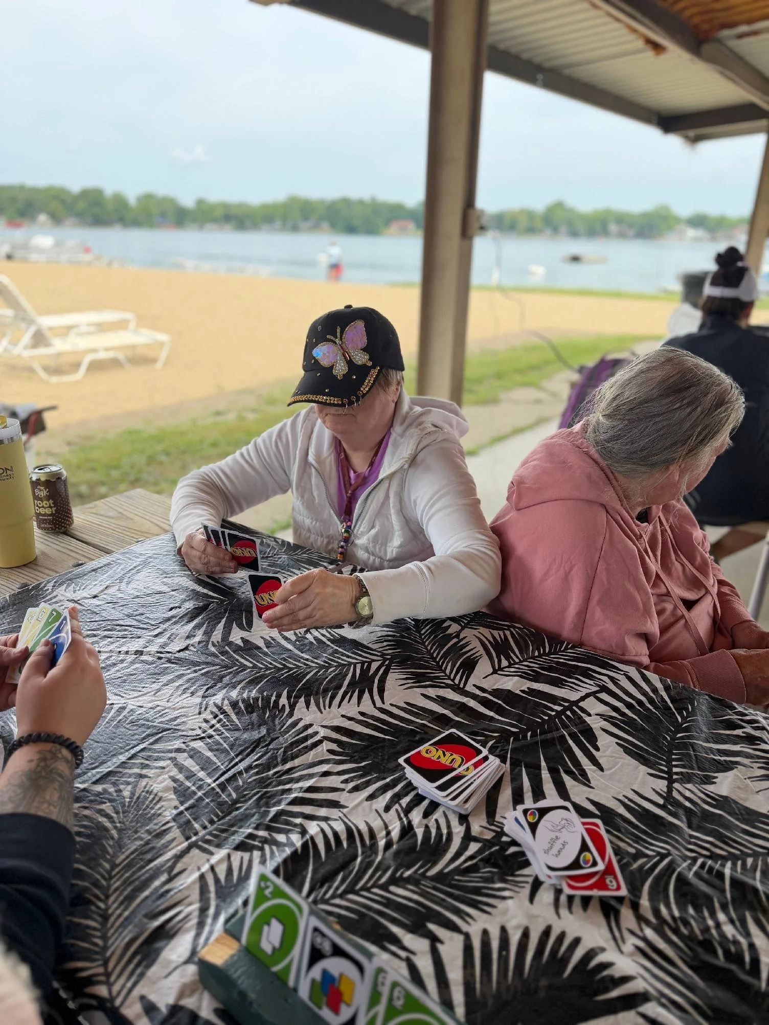 People playing UNO on a table with a tropical leaf patterned tablecloth, outdoors near a lake with boats and trees in the background.