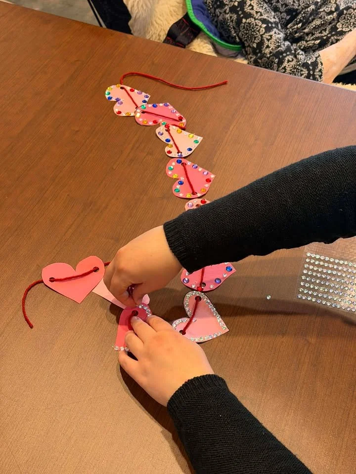 Children are making a heart chain craft with pink and decorated paper hearts connected by a red string on a wooden table.
