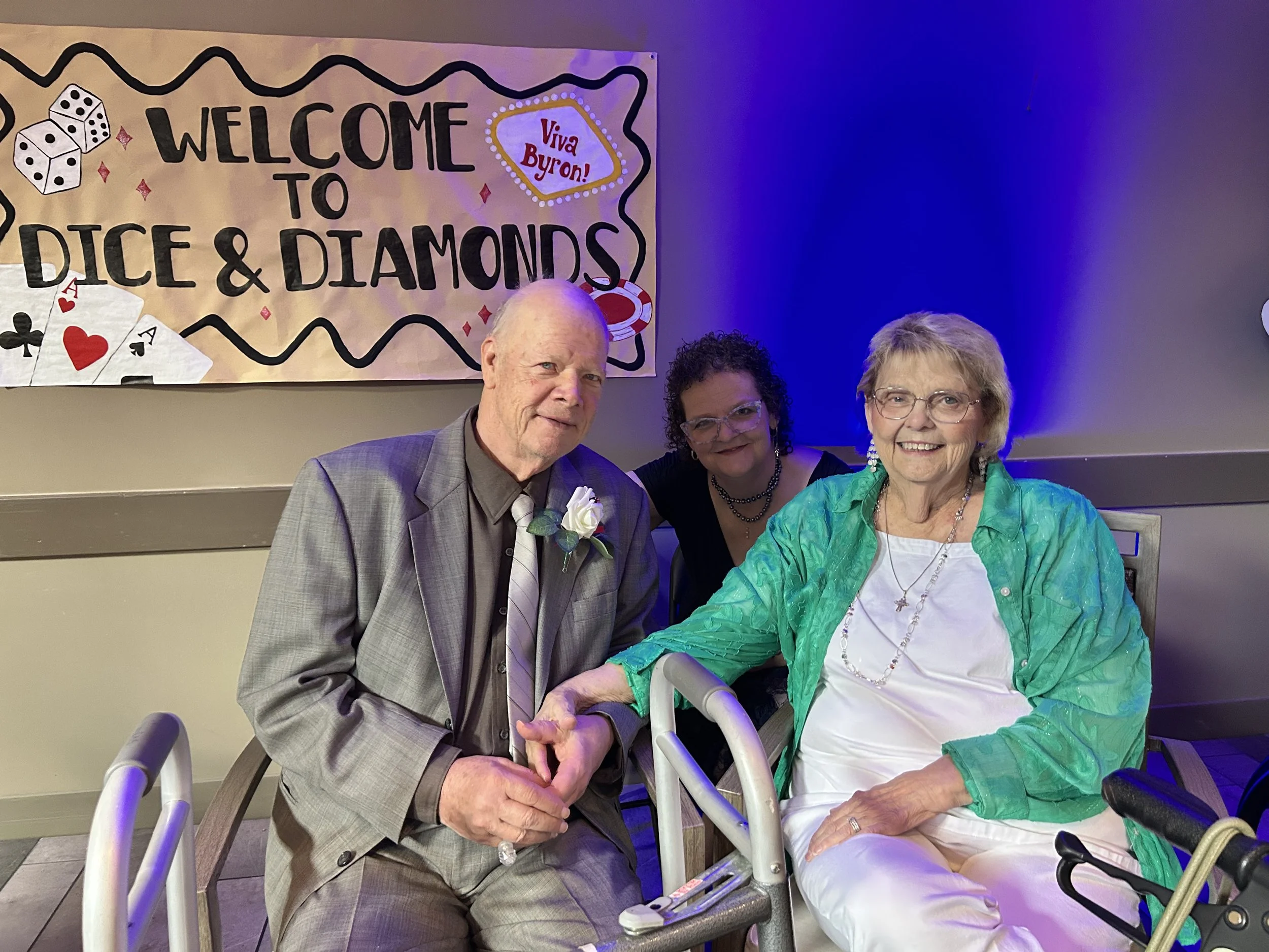 Three older adults, two women and one man, sitting together and smiling at a celebration or gathering. The man is in a gray suit with a boutonniere, and one woman is dressed in a bright turquoise jacket. They are in front of a decorated wall with a b