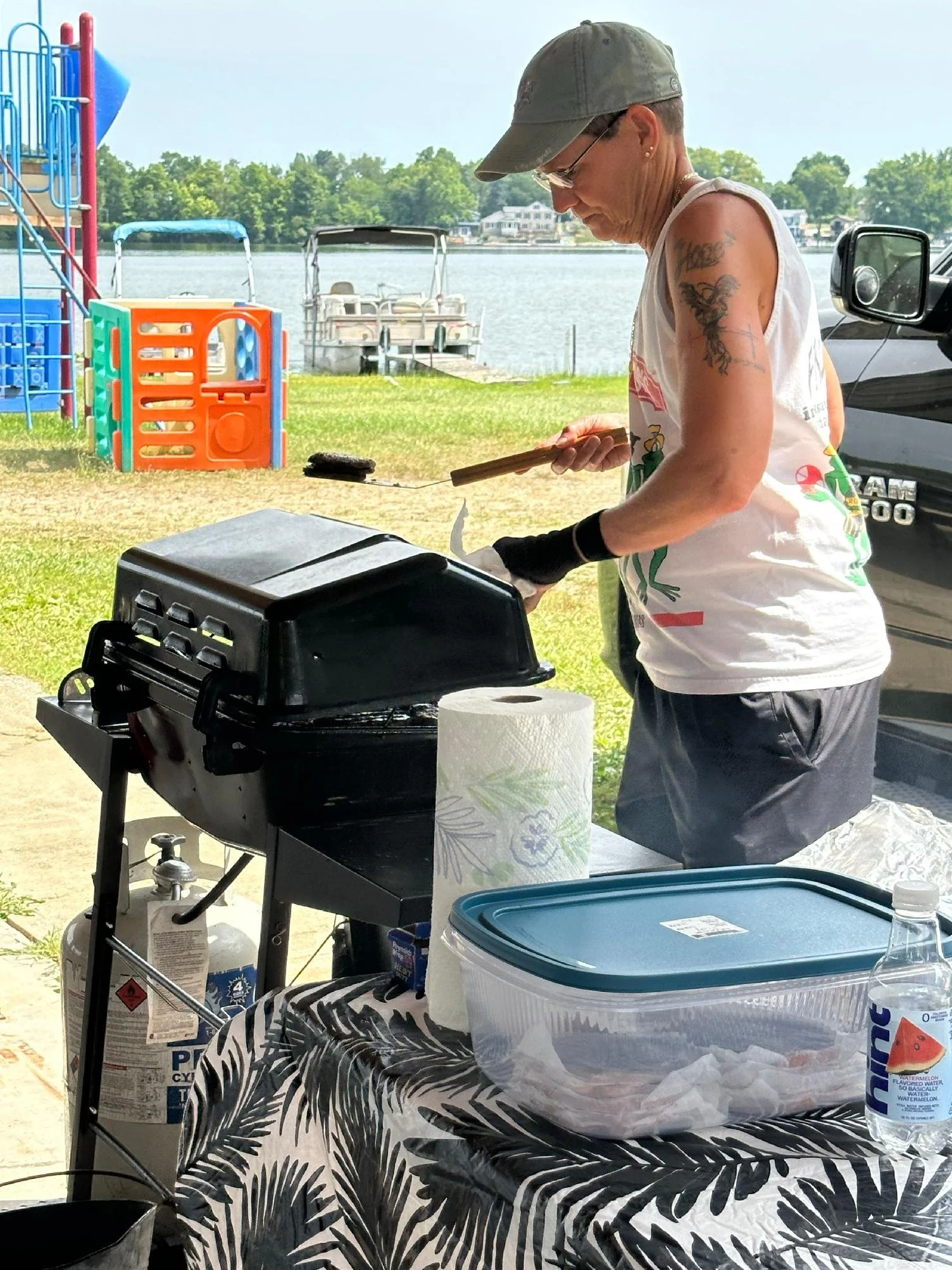 A woman grilling outside near a lake with a boats in the background. She is wearing a gray cap, glasses, and a sleeveless shirt. There are paper towels, a blue container, and other items on a table covered with a black and white leaf-patterned tablec