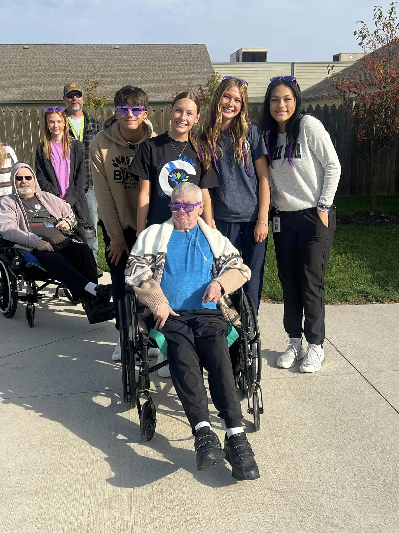 A group of people outdoors on a sidewalk, including a young man in a wheelchair, several young women, and two older men, with wooden fences, trees, and houses in the background.