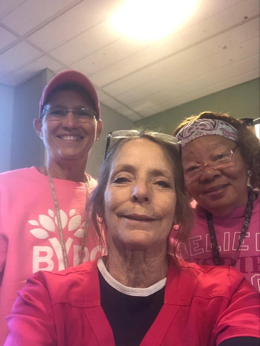 Three women wearing pink clothing taking a close-up selfie indoors, smiling at the camera.
