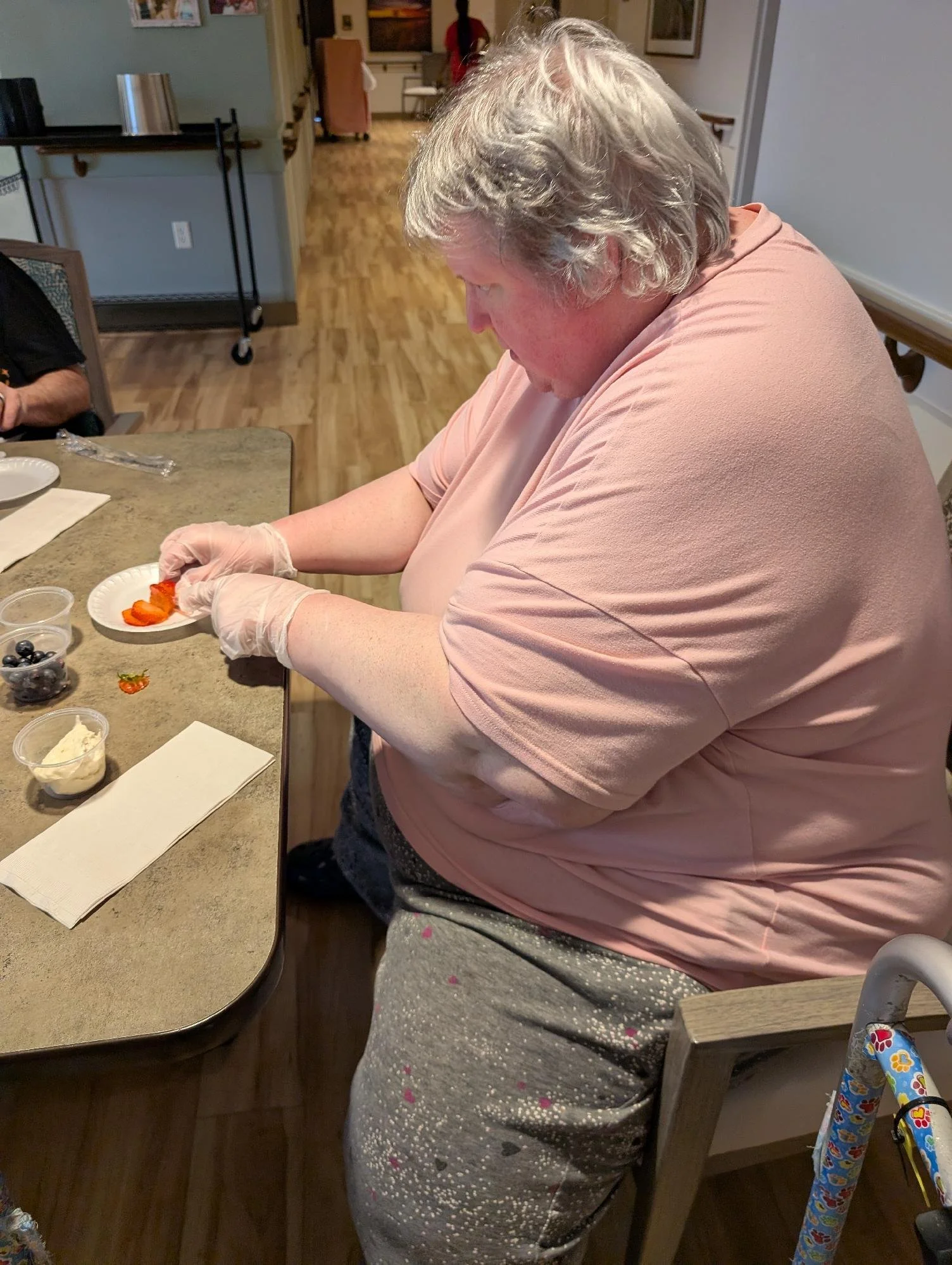 An elderly woman with gray hair is sitting at a kitchen table, wearing a pink shirt and glasses, preparing a meal with sliced strawberries. There are small containers of blueberries and whipped cream on the table, along with utensils, napkins, and a 
