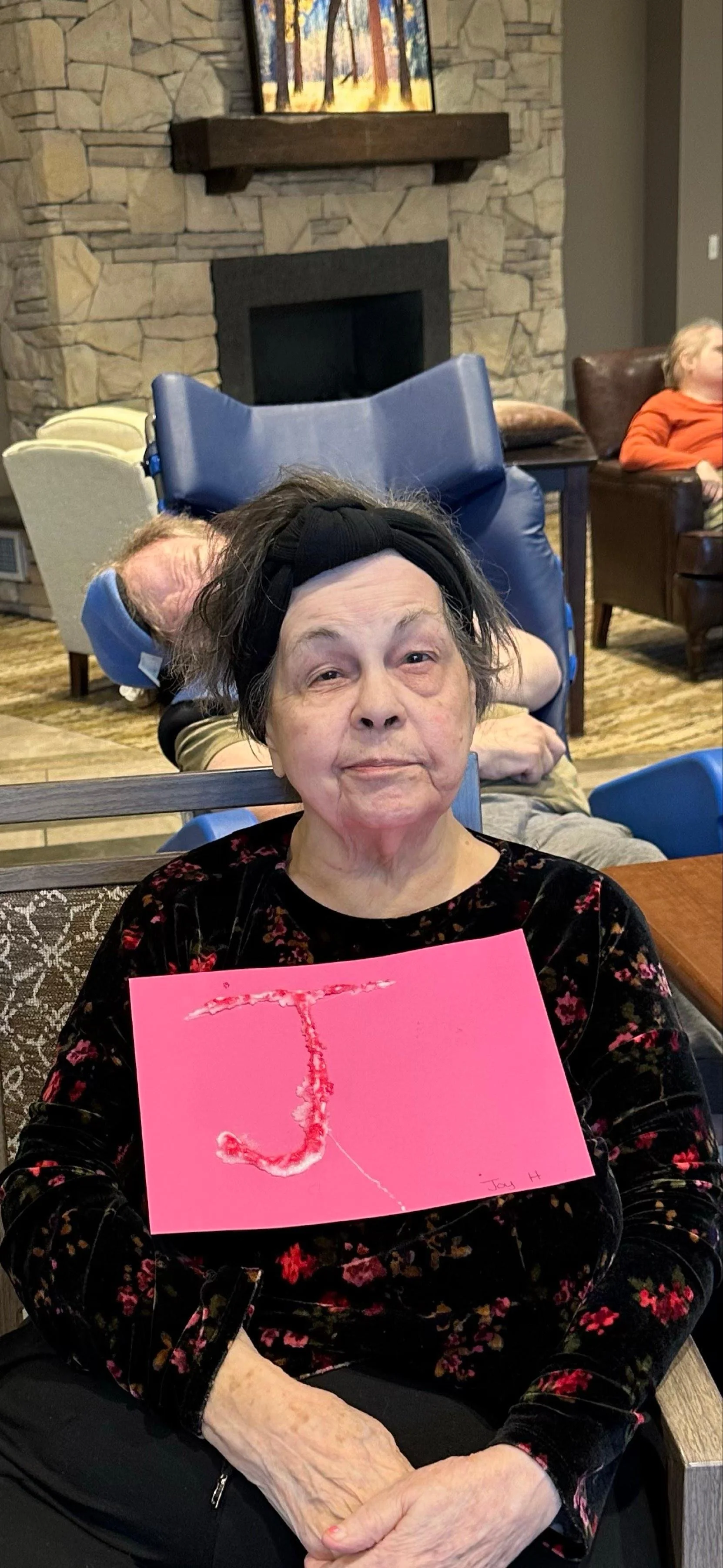 An elderly woman with short gray hair and wearing a black headband sits in a chair, holding a pink card with a red, tree-shaped design and the name "Joy H" written at the bottom right corner. She has a serious expression and is dressed in a black flo