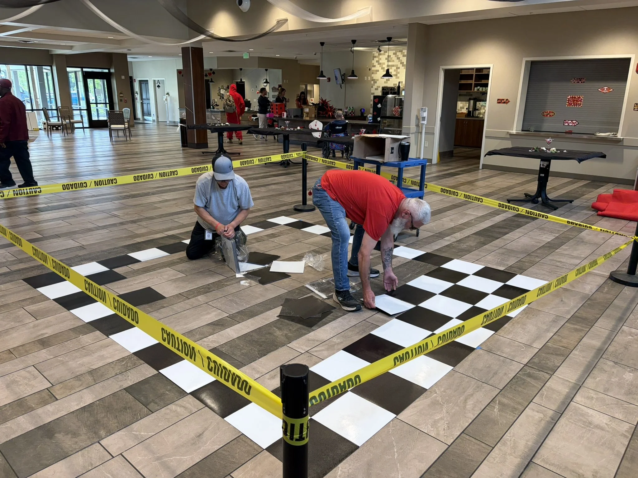 Indoor space with a black and white checkered floor pattern sectioned off by yellow caution tape. Two people are working on the floor, setting tiles or assembly. The background shows a reception or cafe area with tables, chairs, and people standing n