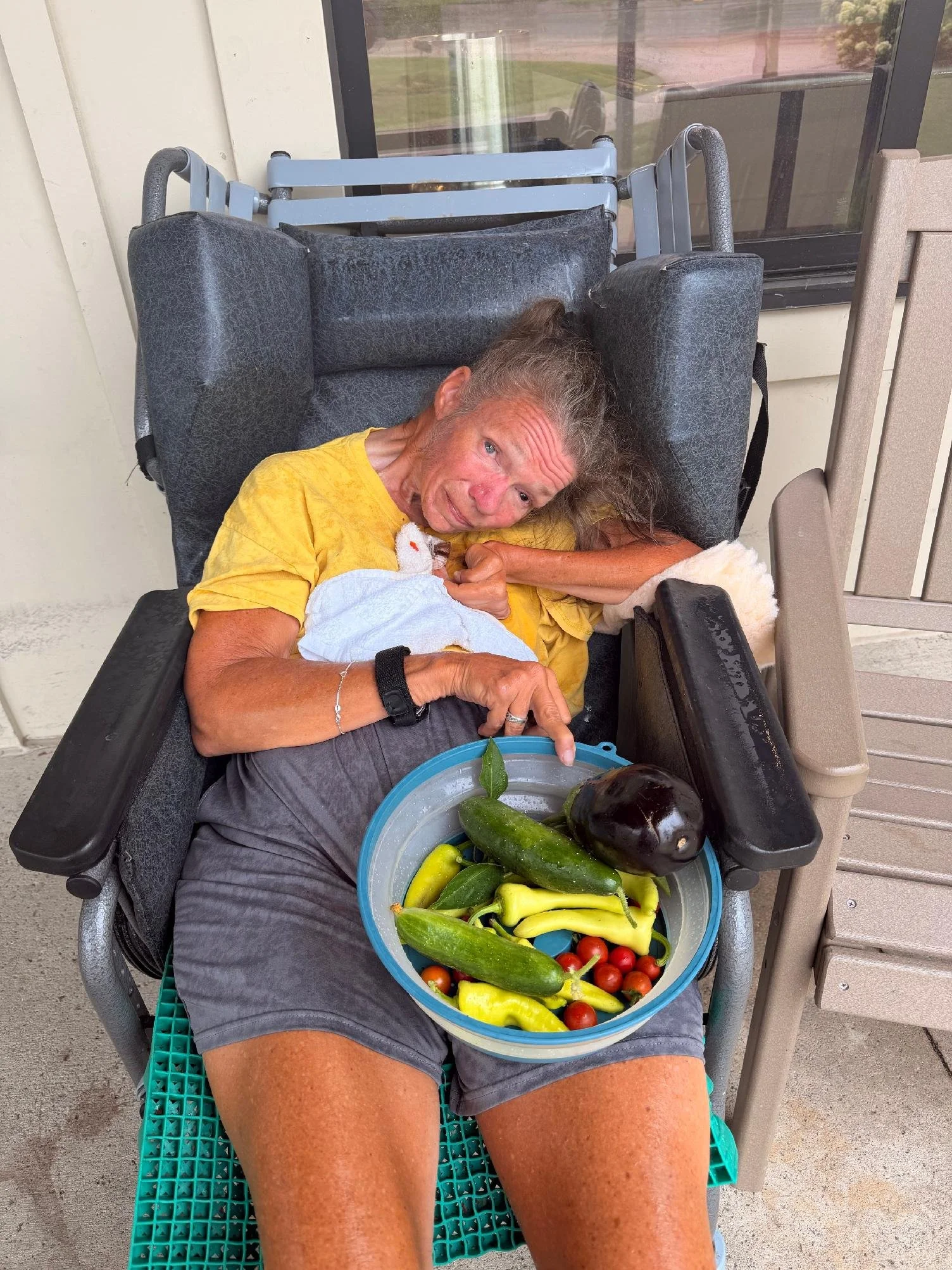 An elderly woman sitting in a wheelchair outdoors, holding a basket of fresh vegetables including zucchini, eggplant, and cherry tomatoes.