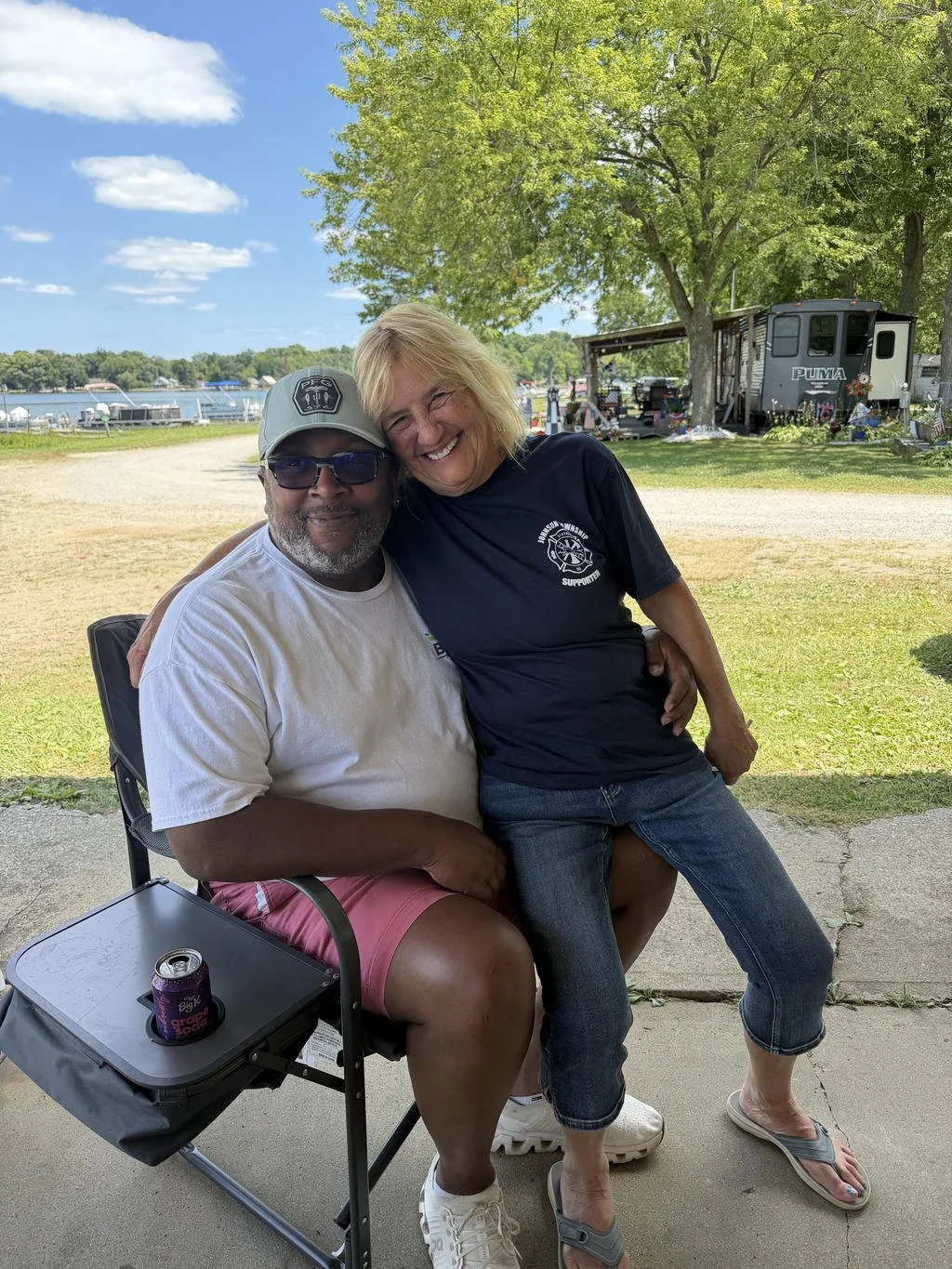 A smiling couple sitting closely together outdoors near a lake, with trees, a trailer, and boats in the background, enjoying a sunny day.