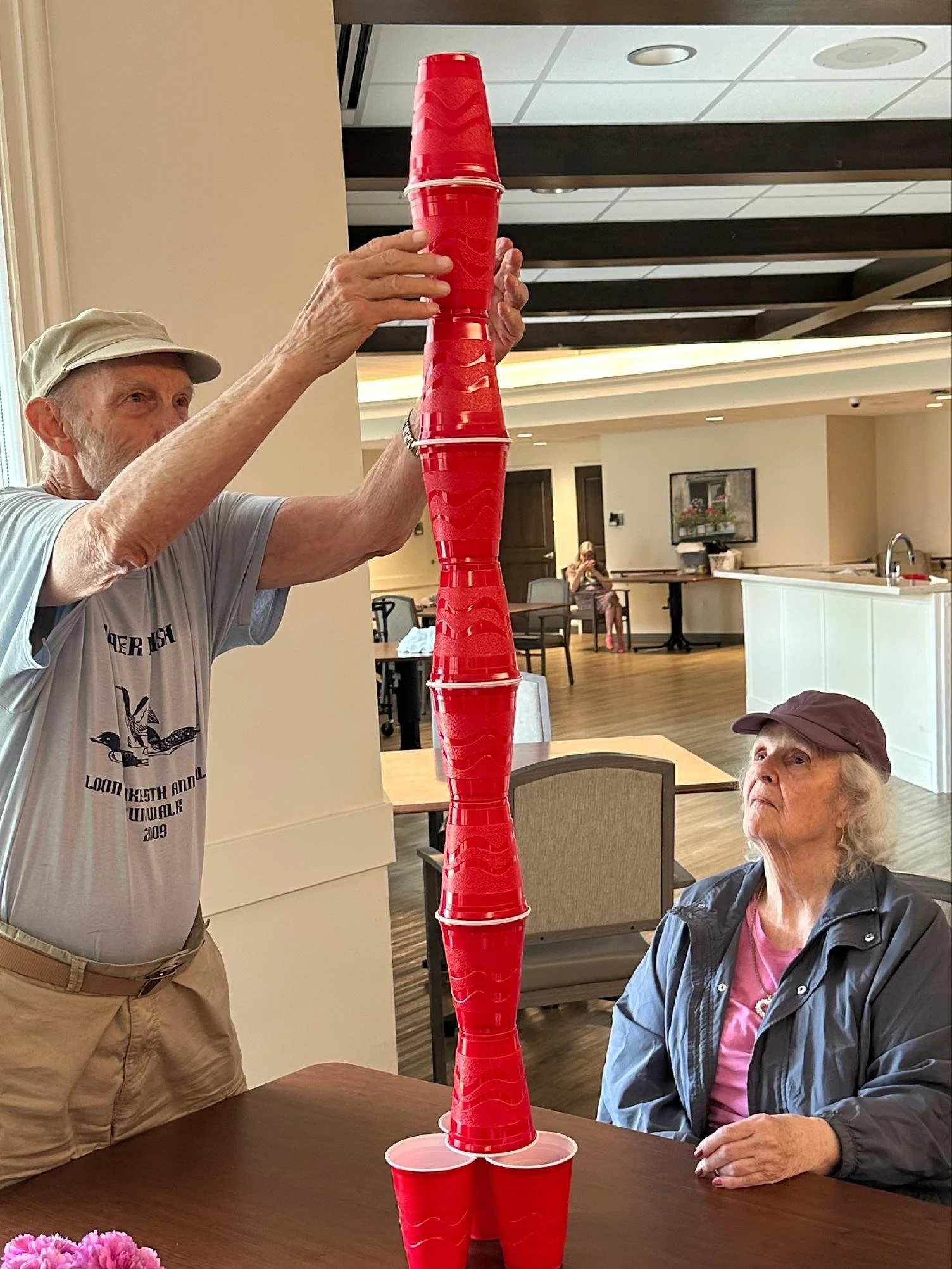 An elderly man and woman playing a stacking game with red plastic cups in a dining area. The man is stacking cups on a table while the woman watches. There is a pink potted flower on the table and a window with sunlight coming through.