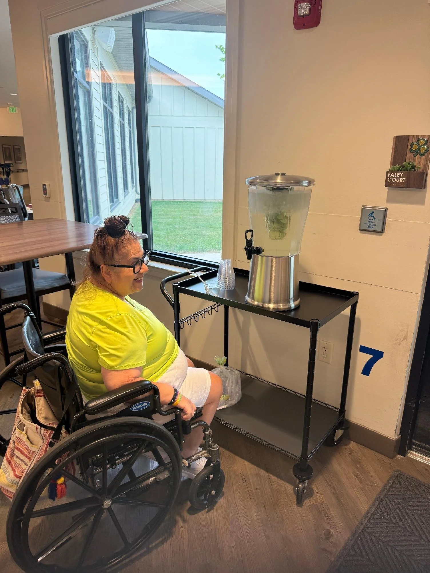A woman in a wheelchair smiling at a water dispenser in a room with wood floors and a large window. The water dispenser has a clear container of water and a silver lid. There is a sign on the wall nearby indicating wheelchair accessibility.