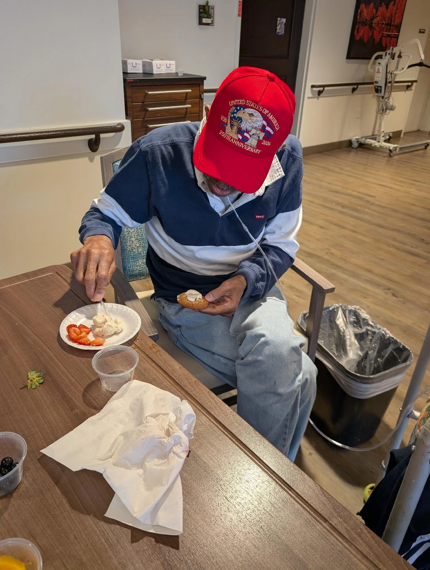 An elderly man wearing a red baseball cap with a patriotic eagle design, and a navy and white jacket, is seated at a wooden table in a dining room of a care facility. He is holding a piece of bread with a spread. The table has a paper plate with stra