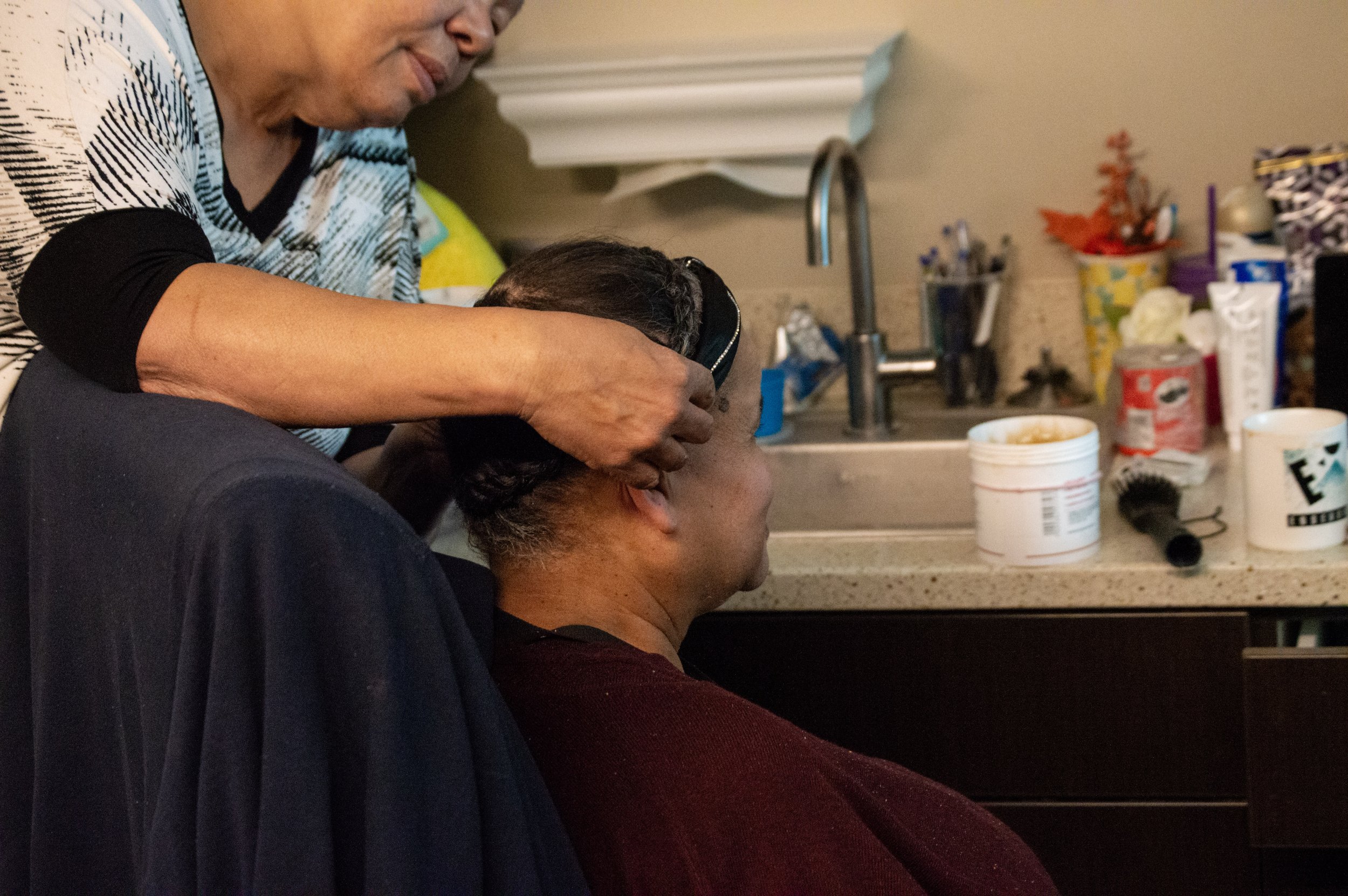 A man with a patterned shirt is washing or fixing the hair of a woman with dark hair, who is seated at a kitchen counter. Various household items, including cups, containers, and decorative items, are visible on the counter.