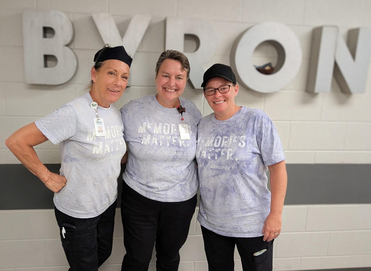 Three women standing together, smiling, in front of a wall with large metallic letters spelling 'BYRON'. They are wearing light-colored T-shirts with the words 'MEMORIES MATTER' and 'End Alzheimer's' printed on them.