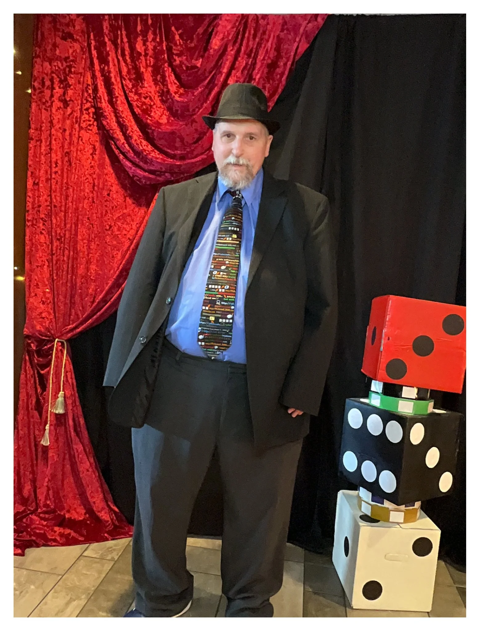 A man dressed in a suit with a colorful patterned tie, gray pants, a blue shirt, and a hat, standing in front of a red velvet curtain, with oversized dice decorations visible on his left side.