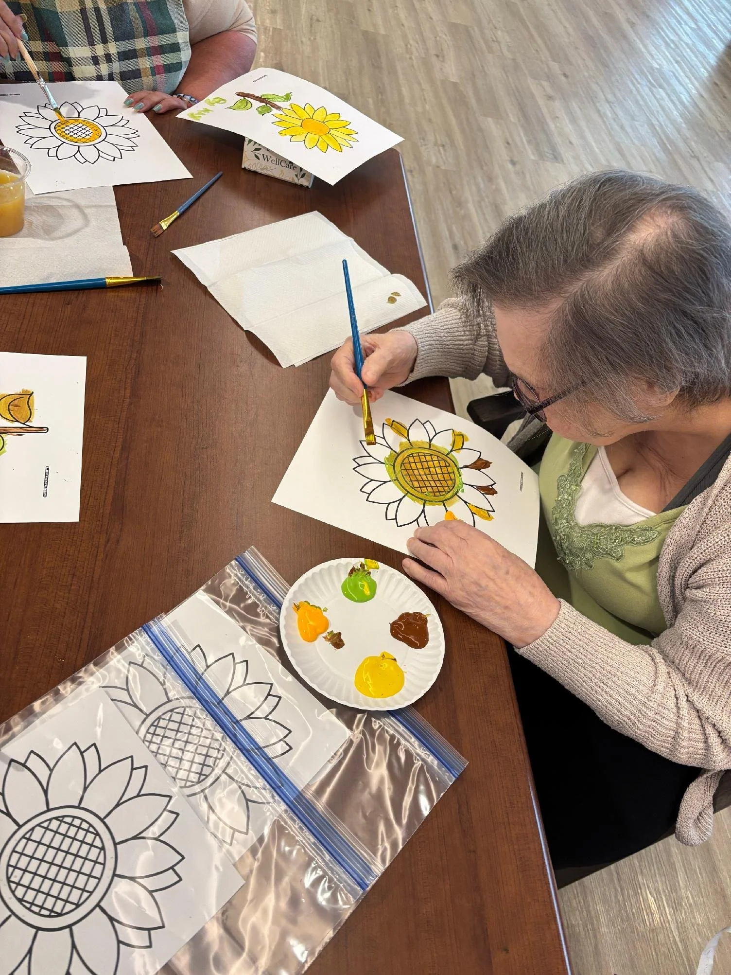 Older woman painting a sunflower on a piece of paper at a table with paint and coloring supplies, with a second person also painting a sunflower in front of her.