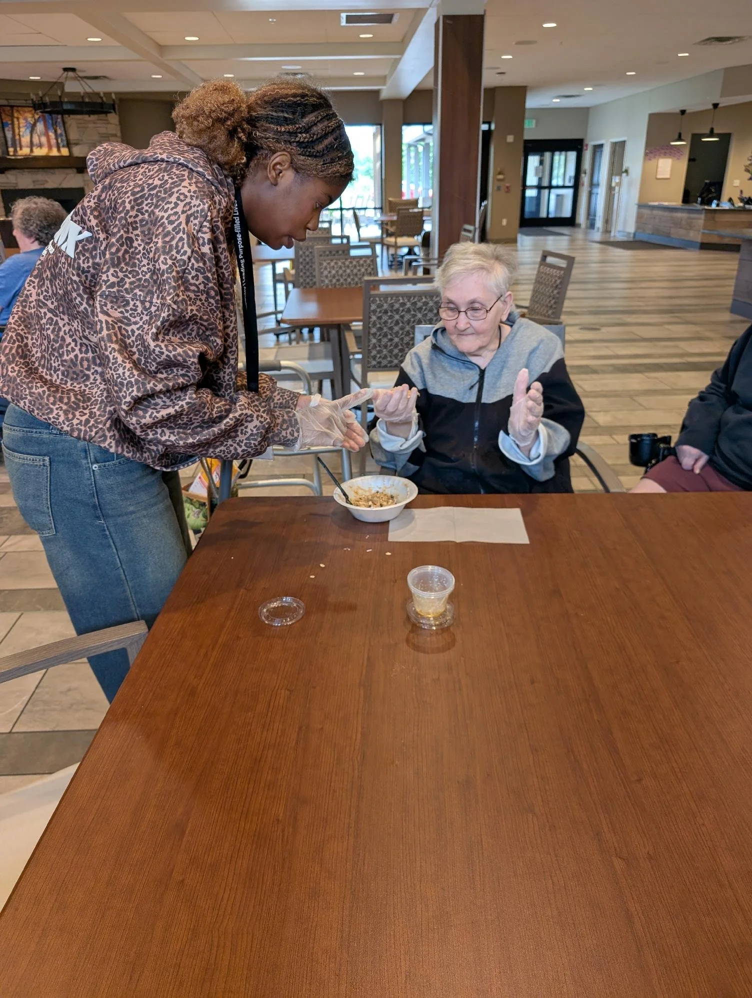 A young woman with braided hair is helping an elderly woman with short gray hair and glasses in a restaurant or cafe. The young woman is wearing a leopard print hoodie and blue jeans, and is leaning over the table to assist the older woman, who is se