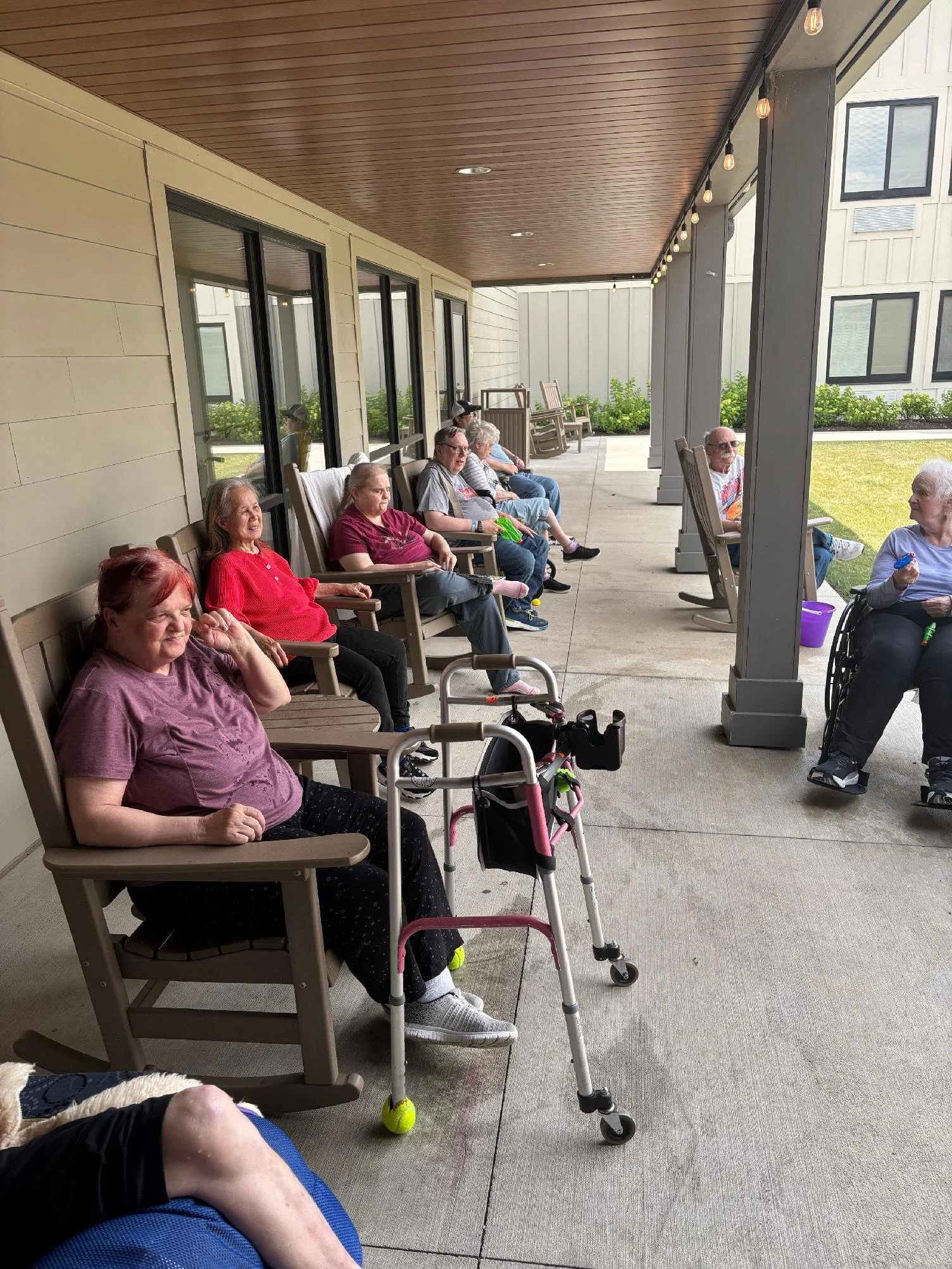 Older adults sitting on a porch, some in chairs, some with walking aids, enjoying the outdoor space with a grassy yard and string lights overhead.