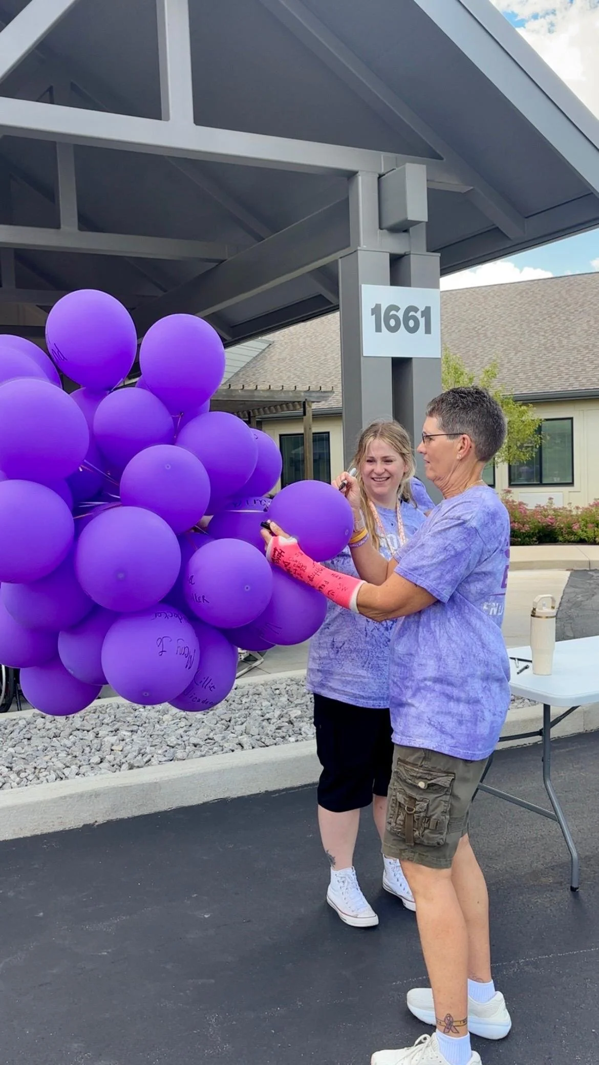 Two women are standing outside at a table with a large bunch of purple balloons. One woman is smiling and appears to be signing a balloon, while the other woman is holding her arm. They are near a building with the number 1661 on it, under a gray str