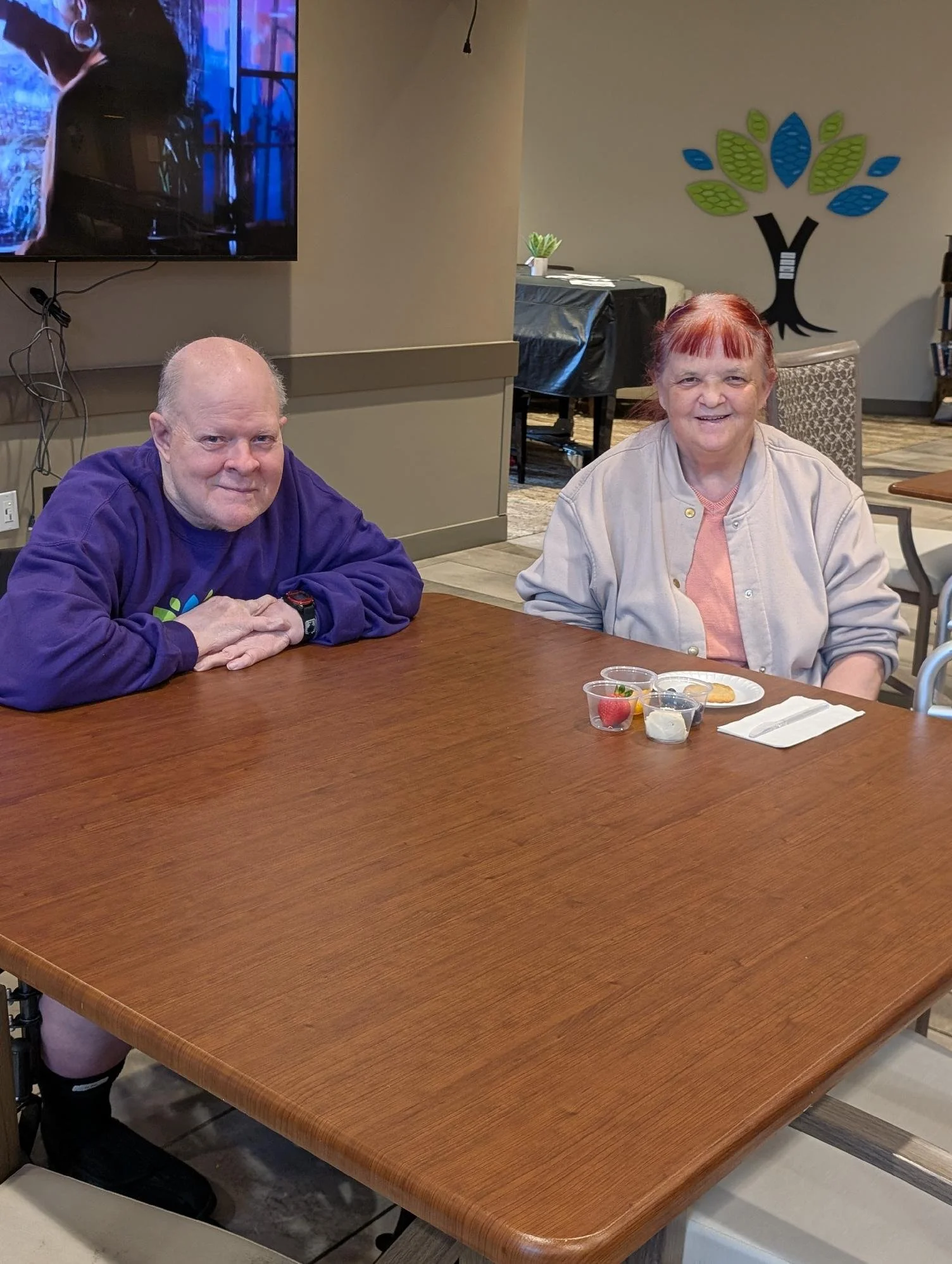 Two older adults sitting at a wooden table, smiling at the camera, with small food containers and utensils in front of them inside a room with a painted tree on the wall.