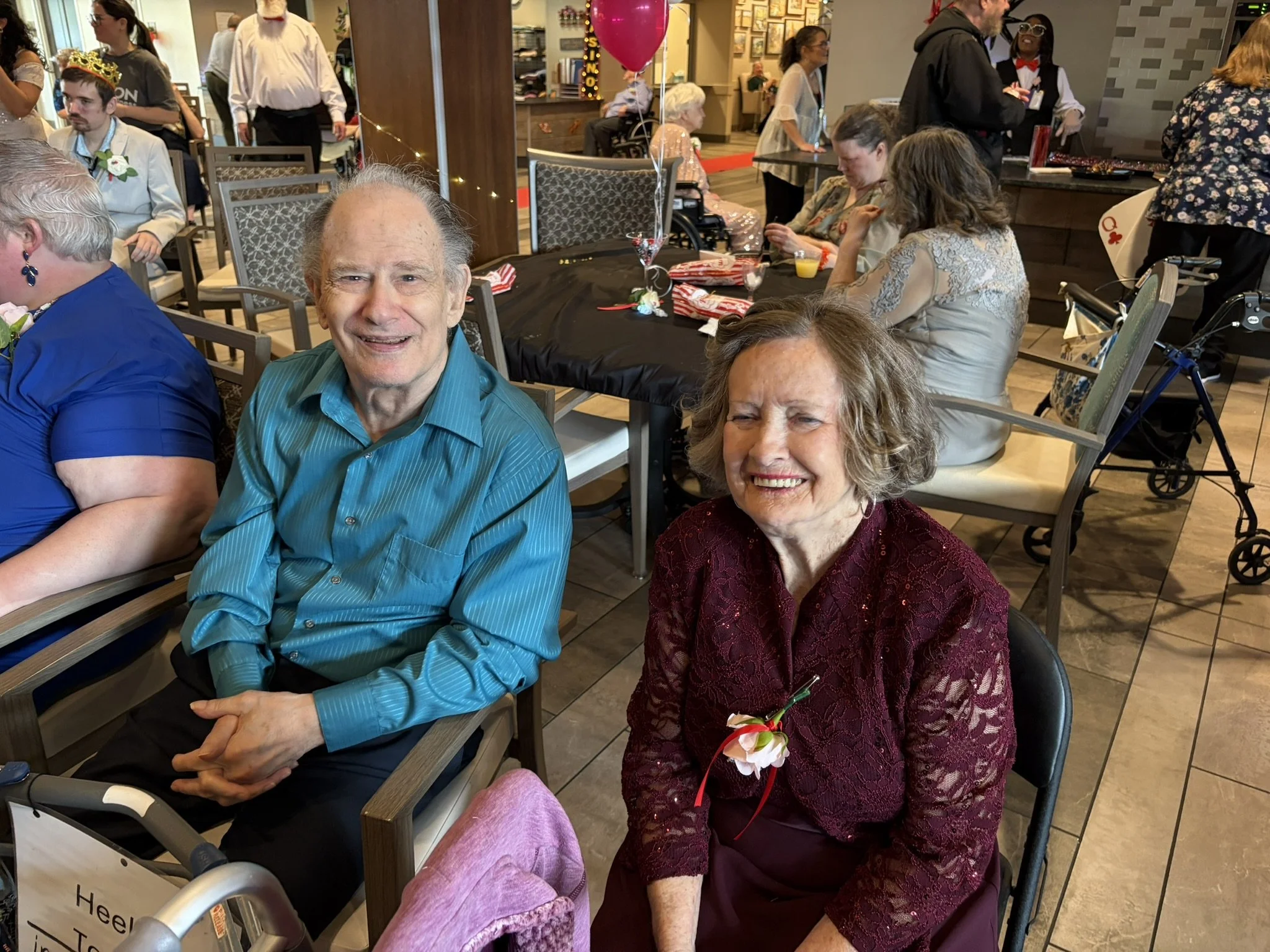 People celebrating at a gathering, with two elderly women sitting in the foreground, one wearing a red lace dress and the other in a blue striped shirt, both smiling. Behind them are other guests seated and standing in a decorated venue, with balloon