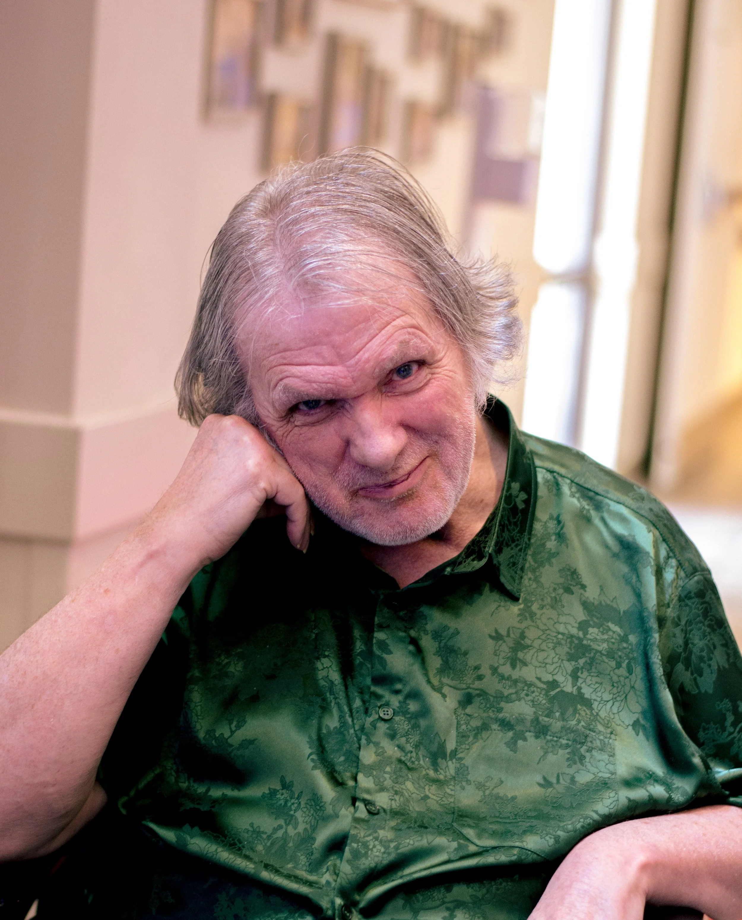 Close-up of an elderly man with gray hair, wearing a green shirt, resting his head on his hand, with a blurred background of framed pictures on a wall.