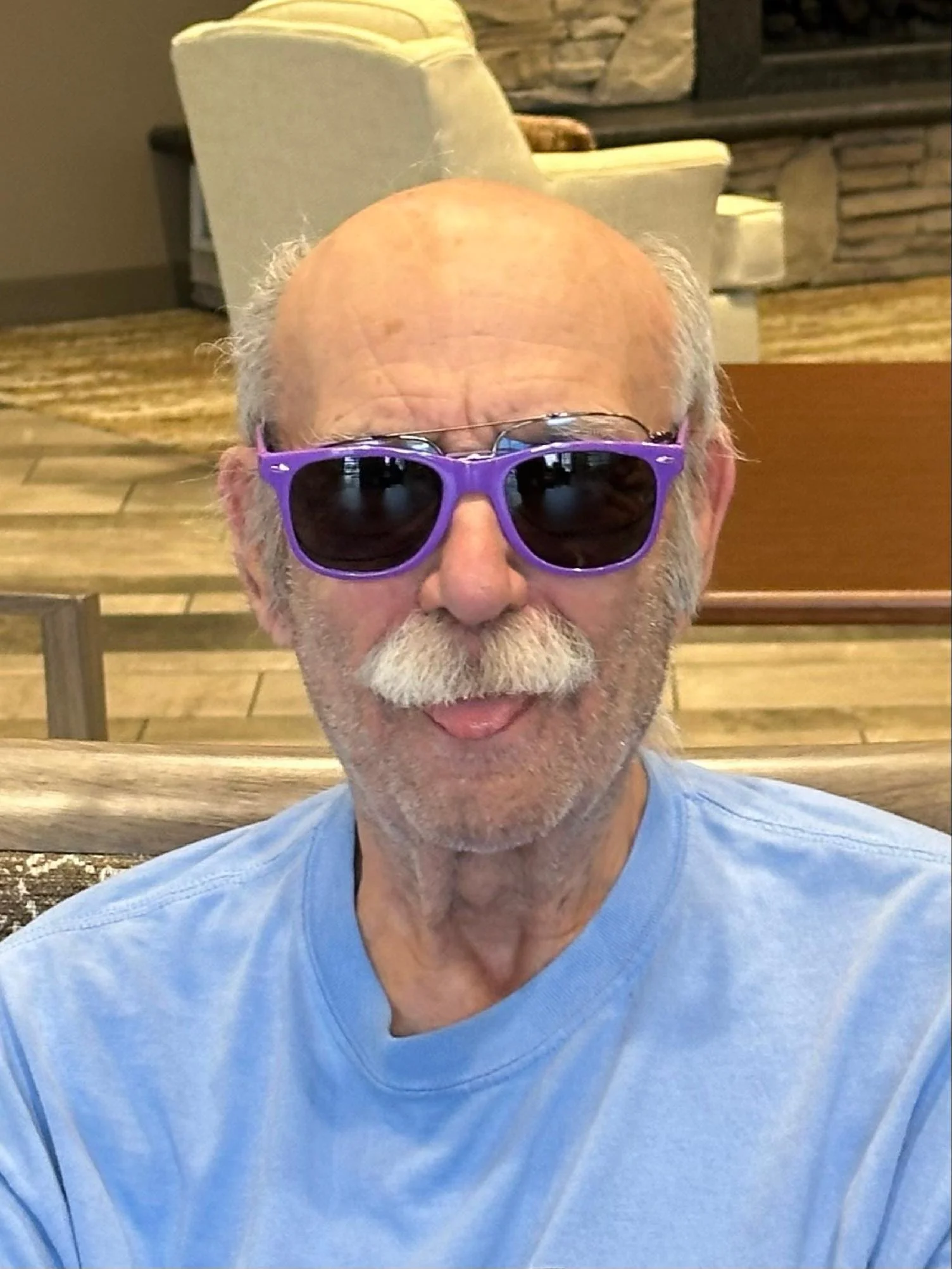 A close-up of an elderly man wearing purple sunglasses and a light blue t-shirt, sitting outdoors with a background showing a stone fireplace and a beige chair.