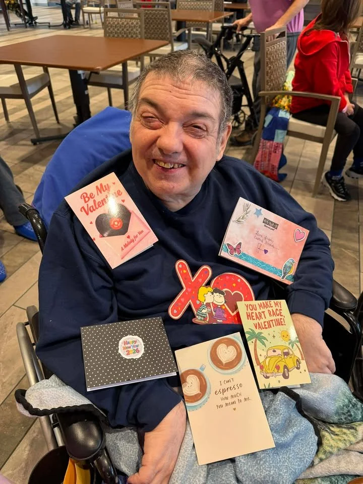 A smiling man sitting in a wheelchair, surrounded by Valentine's Day cards and decorations, in a public indoor space.
