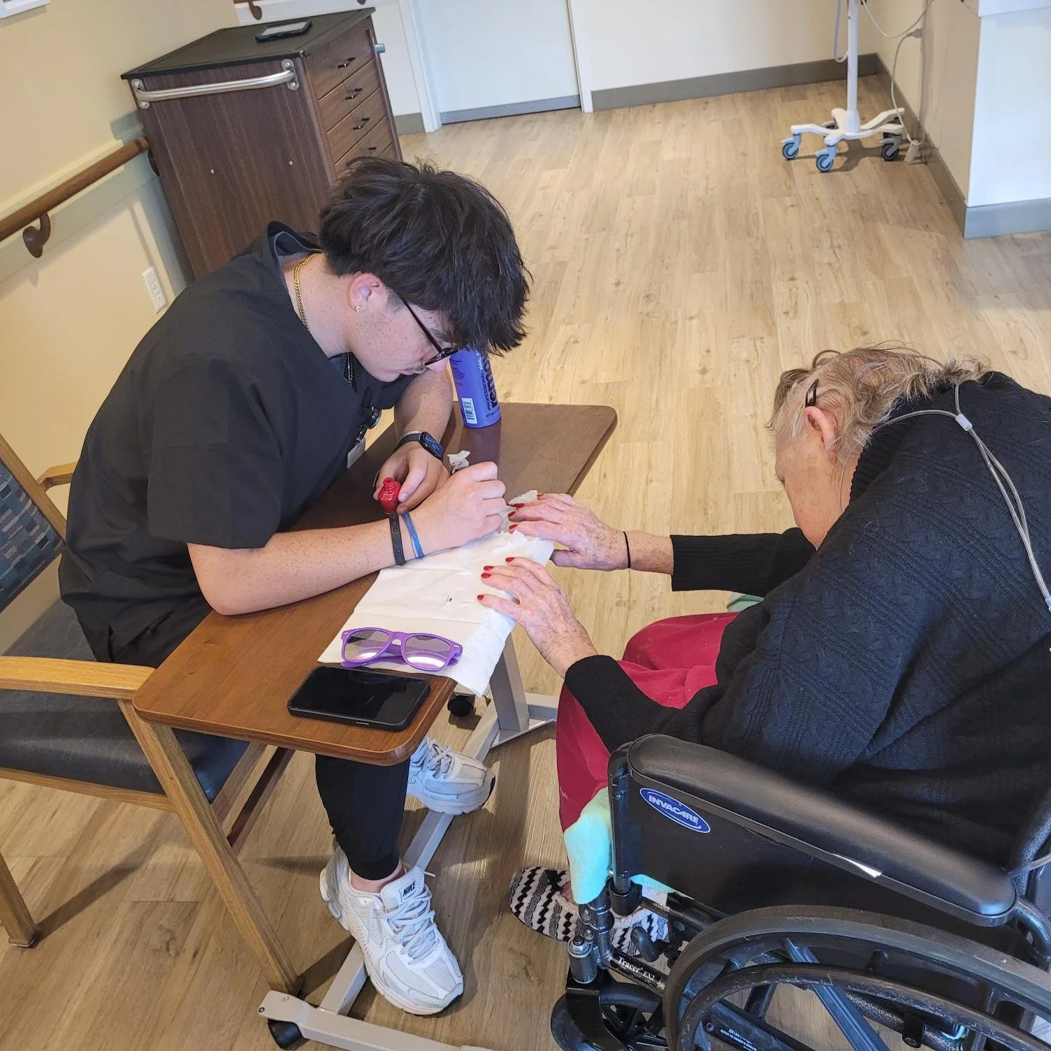 A young male nurse with glasses and tattoos on his arms is drawing on the finger of an elderly woman with light skin, gray curly hair, and glasses, in a healthcare setting. The woman is seated in a wheelchair, and there are personal items like sunglasses, a phone, and a water bottle on the table nearby.
