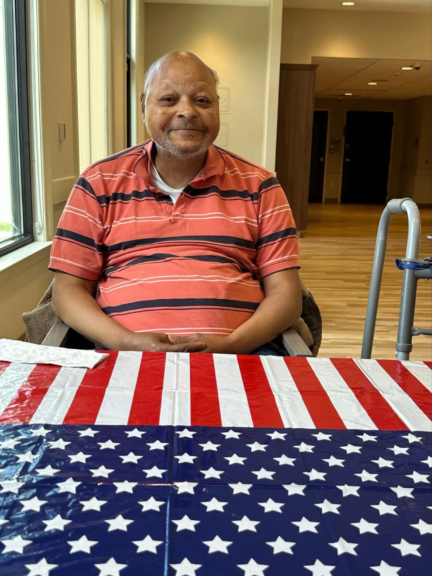 An elderly man sitting at a table covered with an American flag-themed tablecloth, smiling, in a well-lit room with wooden floors and a walker beside him.